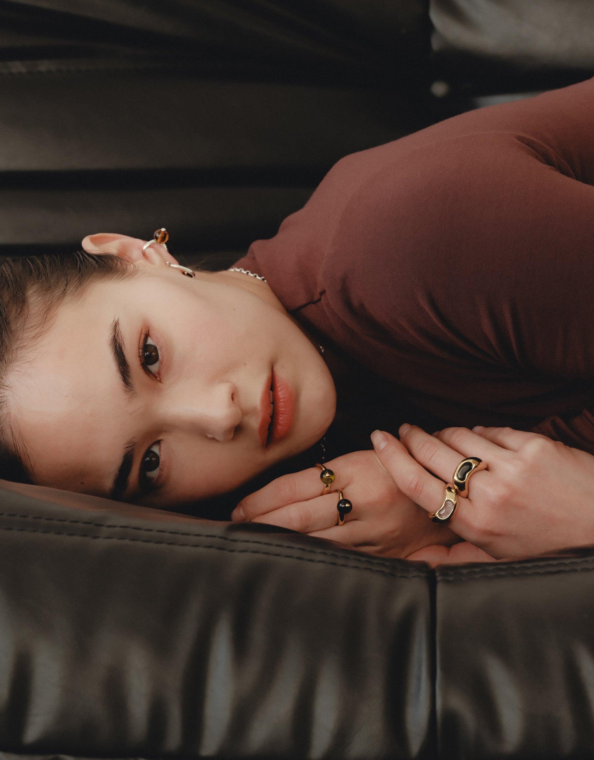 Close-up of a person resting on a leather surface, showcasing multiple rings including the Bean Ring. The jewelry reflects nature-inspired design, emphasizing organic shapes and soft curves reminiscent of legume patterns. CLED Bean Ring.