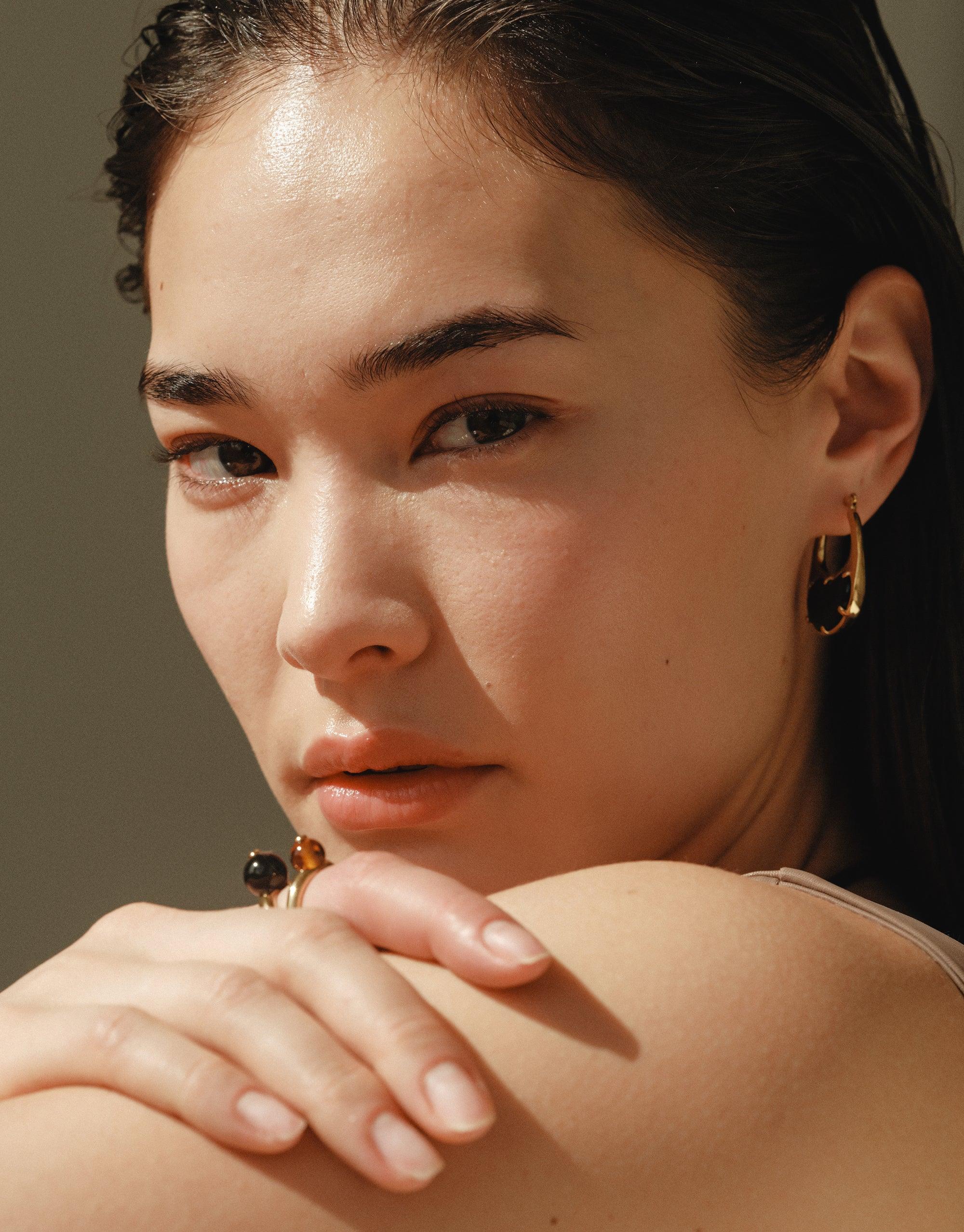 Close-up of a person wearing large gold hoop earrings with a bean-shaped design, showcasing the Bean Hoop Earrings | Large. The earrings elegantly complement the model's features, capturing the organic beauty inspired by nature's legumes. CLED