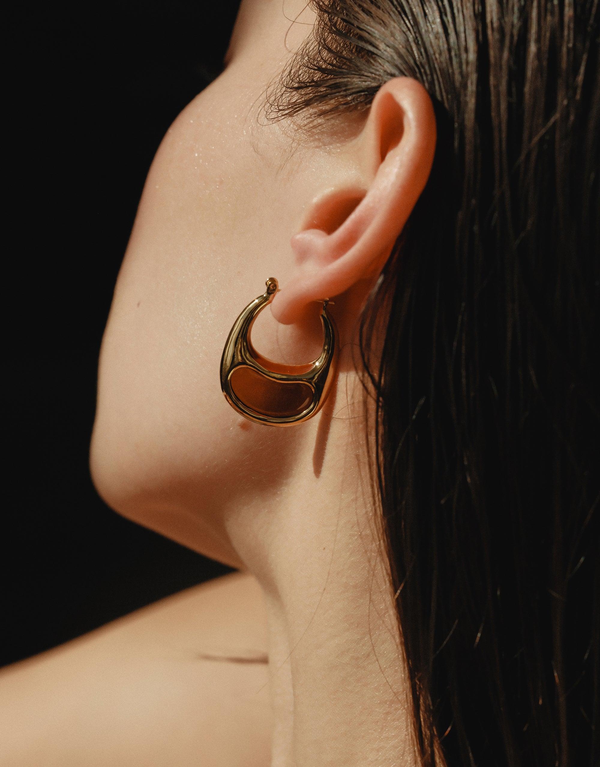 Close-up of a person wearing a large, golden bean-shaped hoop earring, showcasing the organic curves and natural inspiration of the Bean Hoop Earrings | Large. The earring's unique design reflects nature's beauty and creativity. CLED