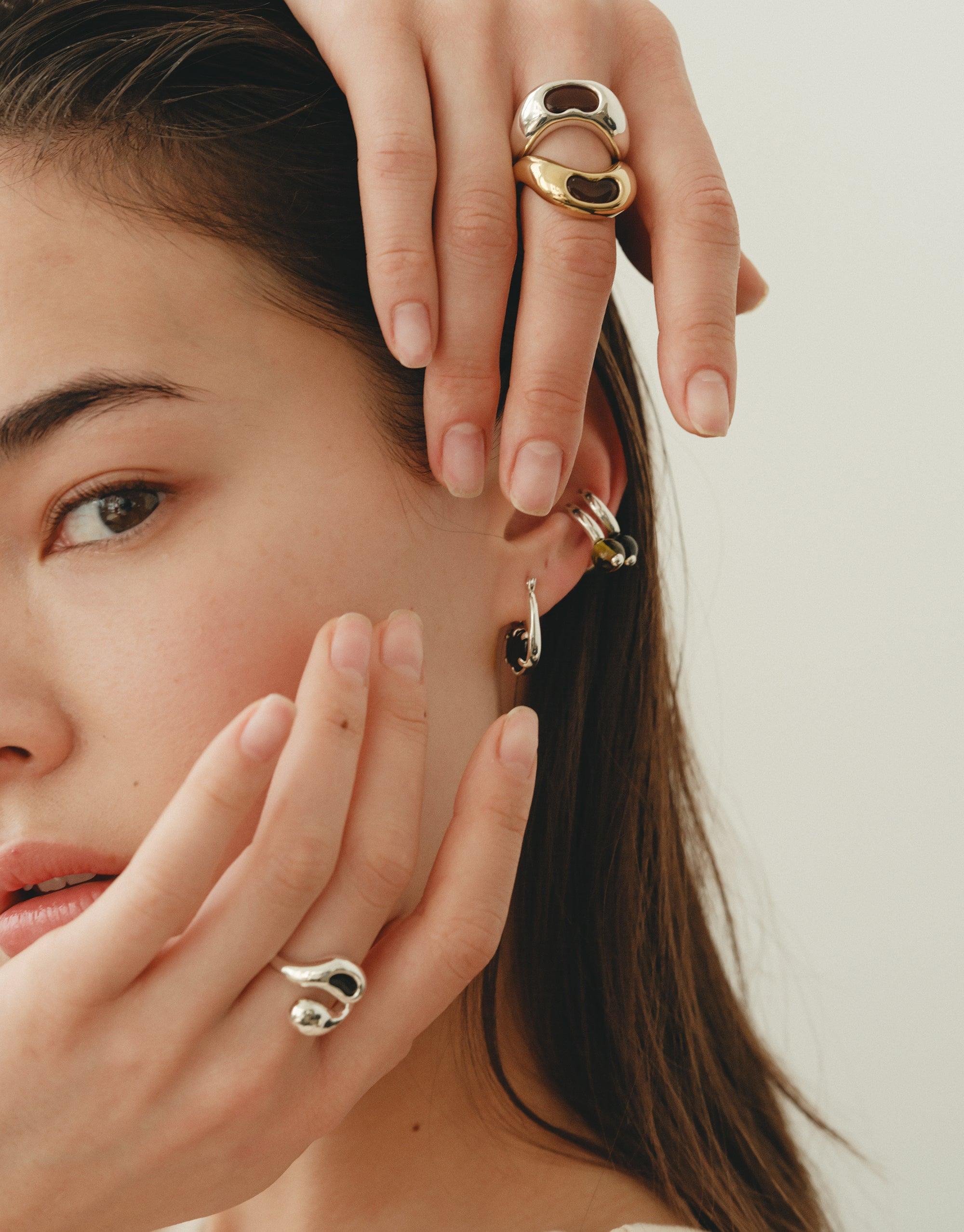 Close-up of hands adorned with elegant Bean Rings, showcasing organic shapes inspired by nature. Earrings complement the rings, highlighting the collection's versatile design. Natural beauty and creativity reflected in jewelry. Bean Ring by CLED.