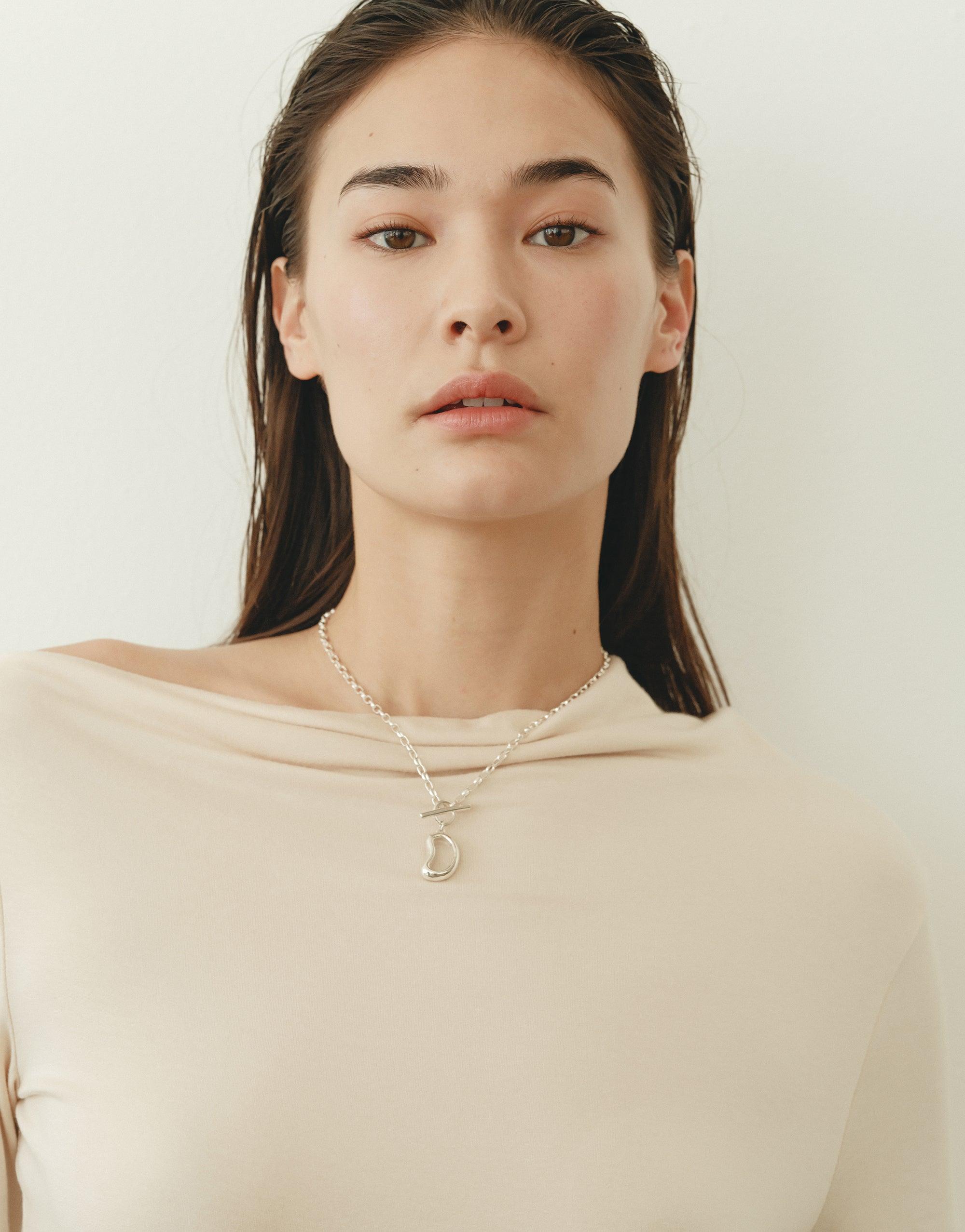 Woman wearing a beige top showcasing a delicate silver necklace with an organic bean-shaped pendant, highlighting the Bean Toggle Necklace's natural and transformative design inspired by legume patterns. The necklace embodies growth and nature's creativity. CLED