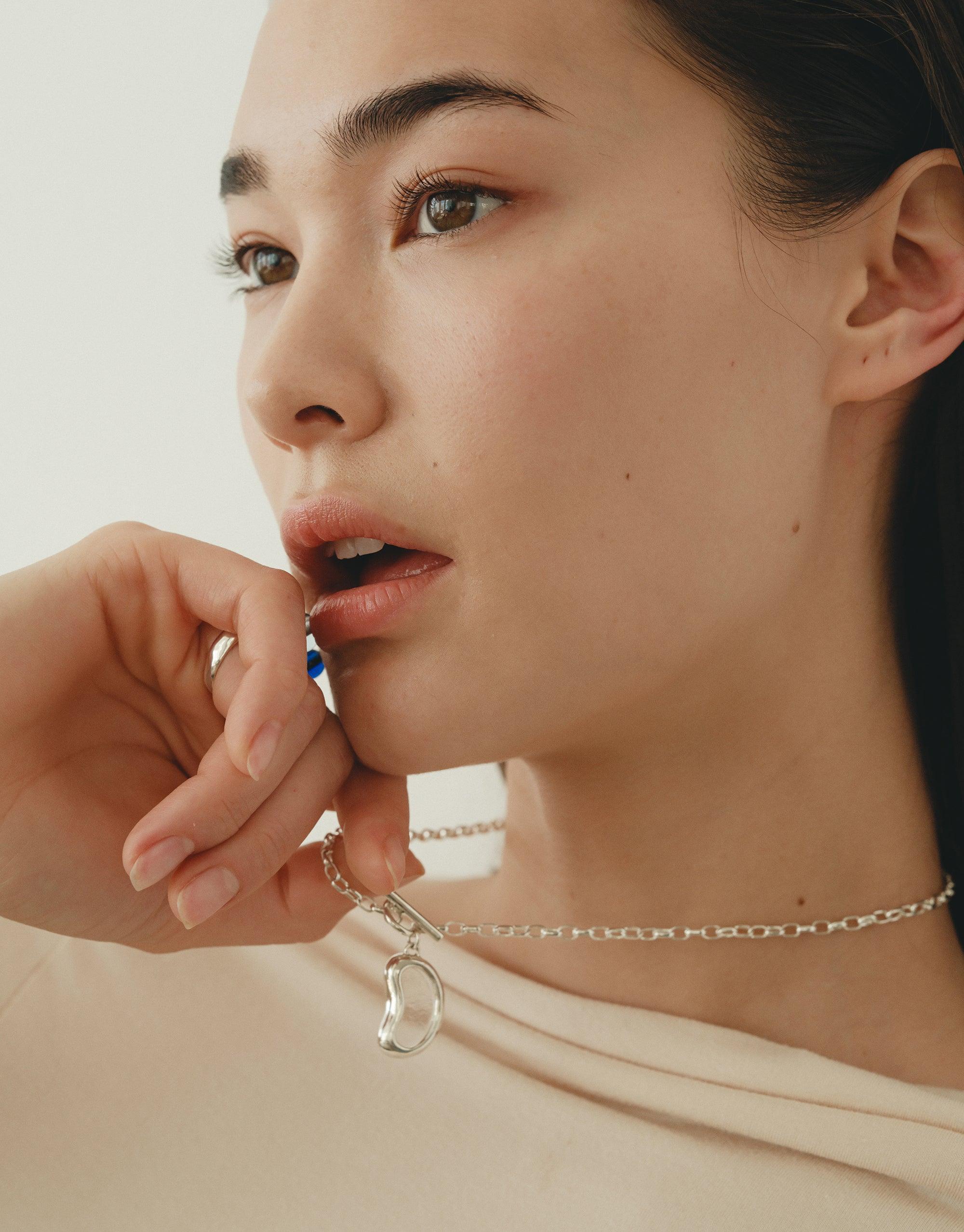 Close-up of a woman wearing a delicate silver chain necklace with a unique bean-shaped pendant, showcasing the Bean Toggle Necklace's elegant design inspired by nature's organic forms. The necklace complements her thoughtful expression, embodying growth and transformation. CLED