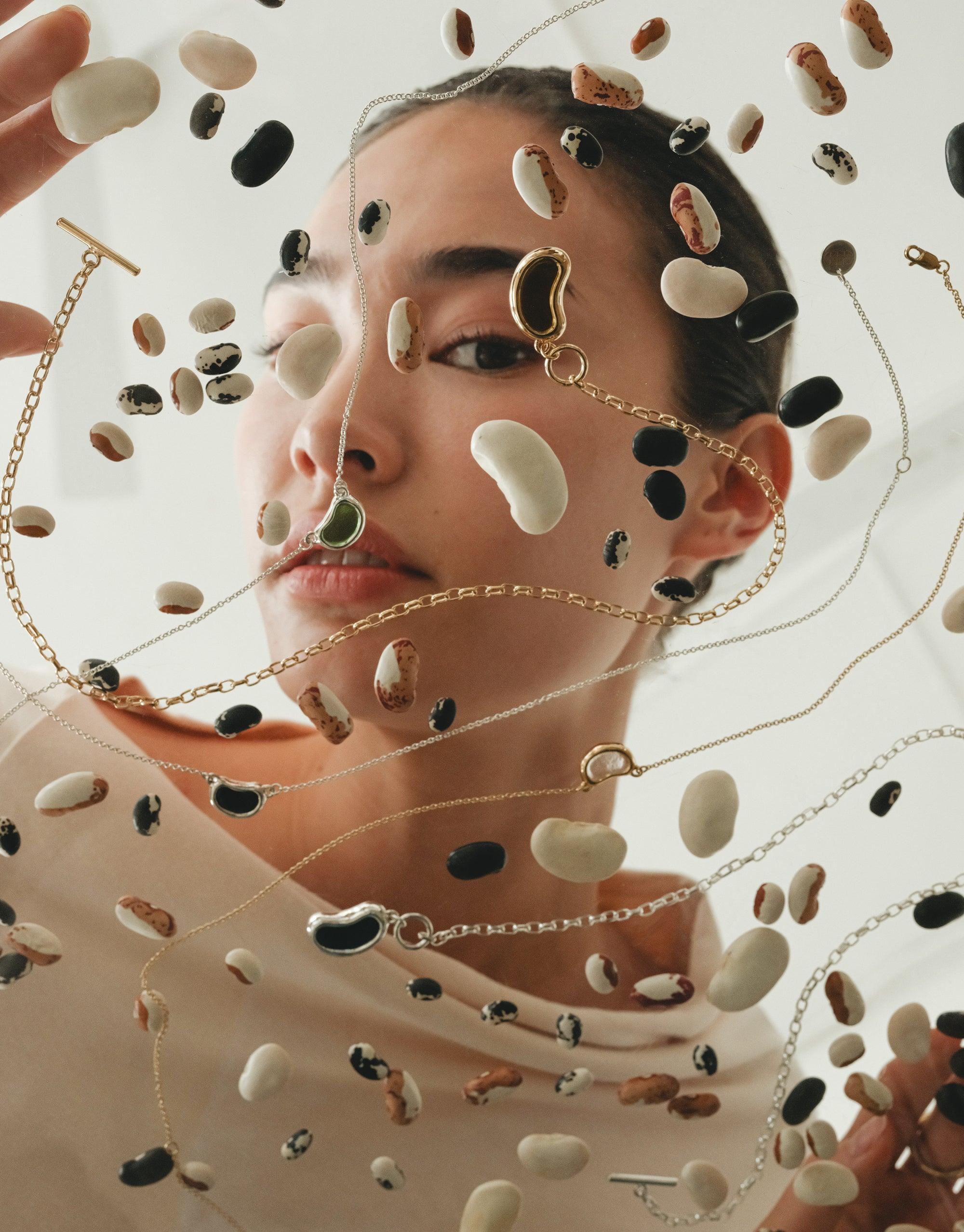 Artistic portrait showcasing Bean Toggle Necklace surrounded by floating legumes and chain jewelry, capturing nature-inspired design. Model's face partially obscured by beans, emphasizing organic shapes and natural beauty. Unique product display for CLED's Bean Toggle Necklace.
