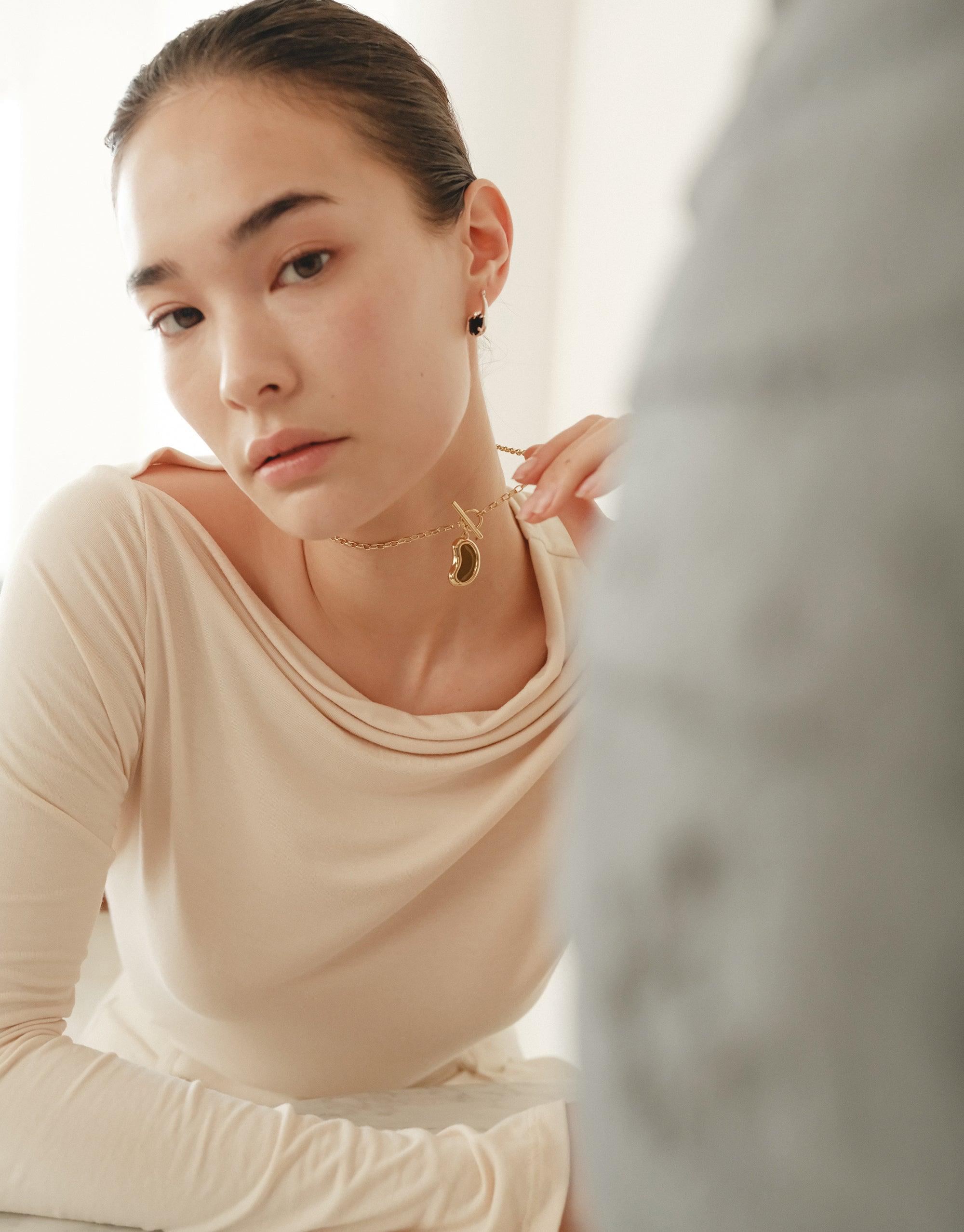 Elegant woman wearing Bean Toggle Necklace, showcasing delicate gold pendant inspired by nature's organic shapes. Soft light enhances the necklace's subtle curves, complementing the model's graceful pose and cream-colored top. Bean Toggle Necklace by CLED.