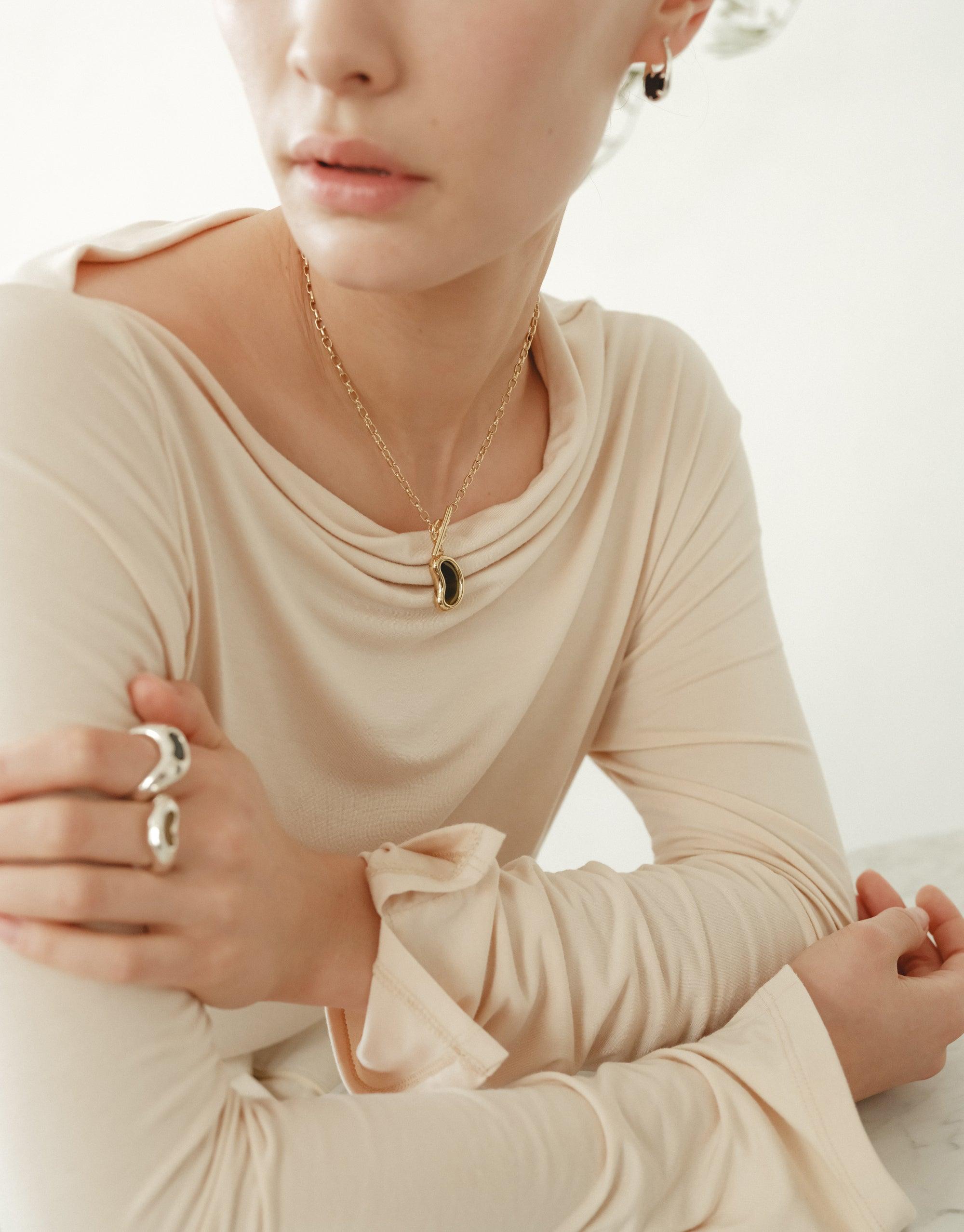 Elegant gold Bean Toggle Necklace featuring a delicate bean-shaped pendant, inspired by nature's organic forms. Worn by a model in a cream-colored long-sleeve top, showcasing the necklace's versatile and sophisticated design. Perfect for adding a touch of natural beauty to any outfit. Bean Toggle Necklace by CLED.