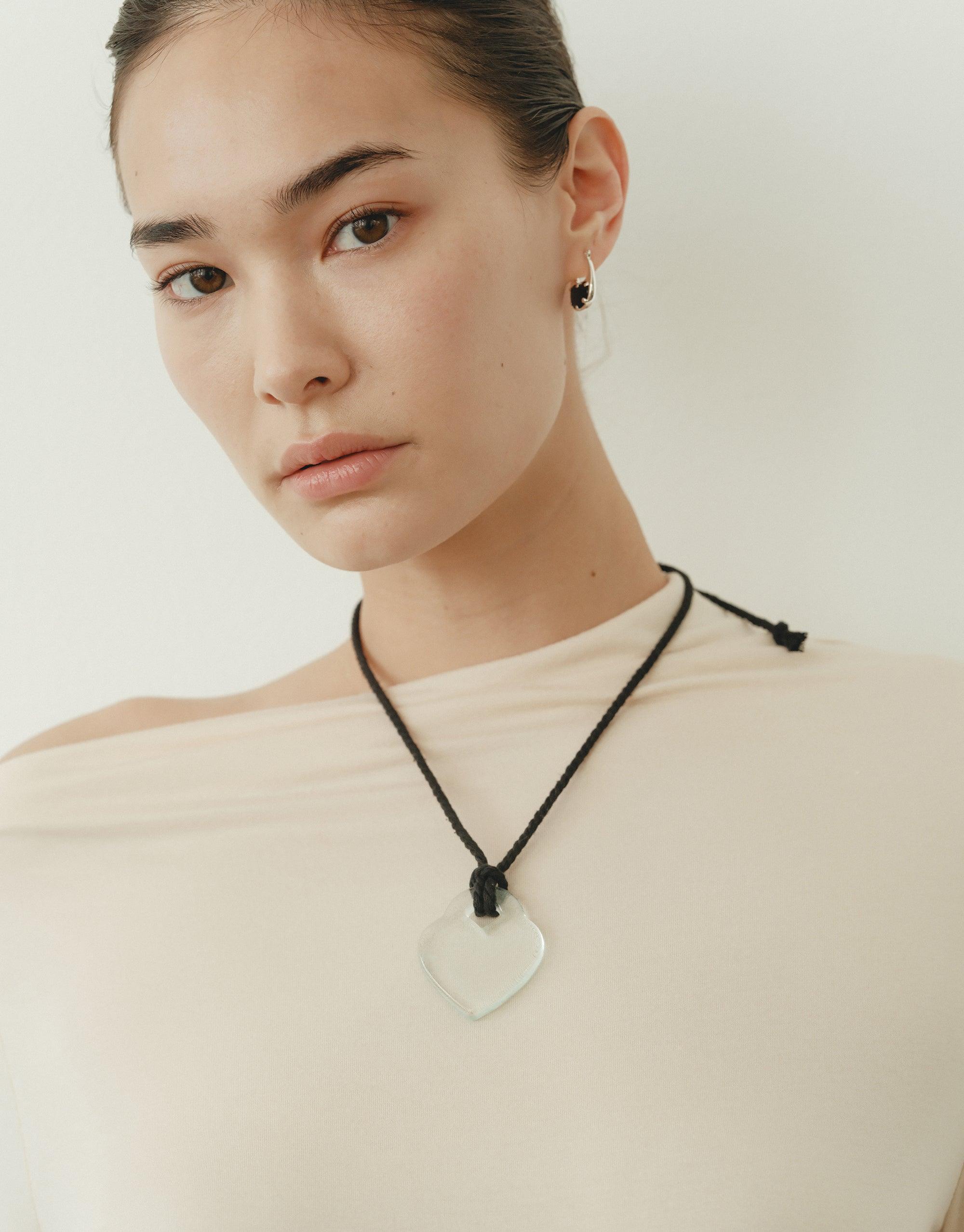 Elegant model wearing Clear Heart Necklace on black cord, showcasing delicate heart-shaped pendant against cream-colored top. Soft, natural lighting emphasizes the necklace's simplicity and beauty. Inspired by organic shapes and nature's creativity. Clear Heart Necklace by CLED.