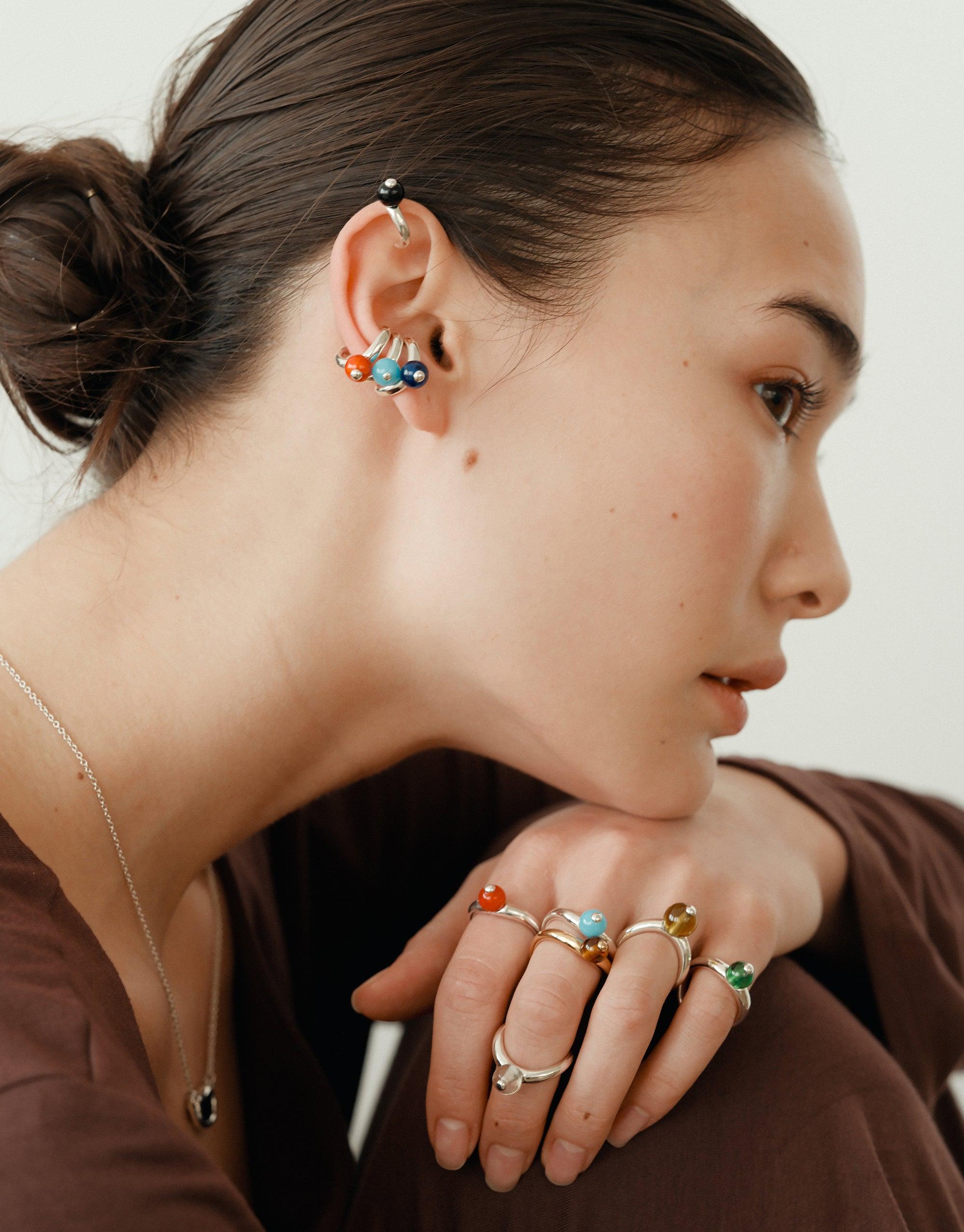 Close-up profile of a woman wearing colorful jewelry, including multiple rings and earrings inspired by organic shapes. Her hair is in a bun, showcasing the unique ear adornments. The image highlights the Pea Curve Ring | Large among other nature-inspired pieces. CLED