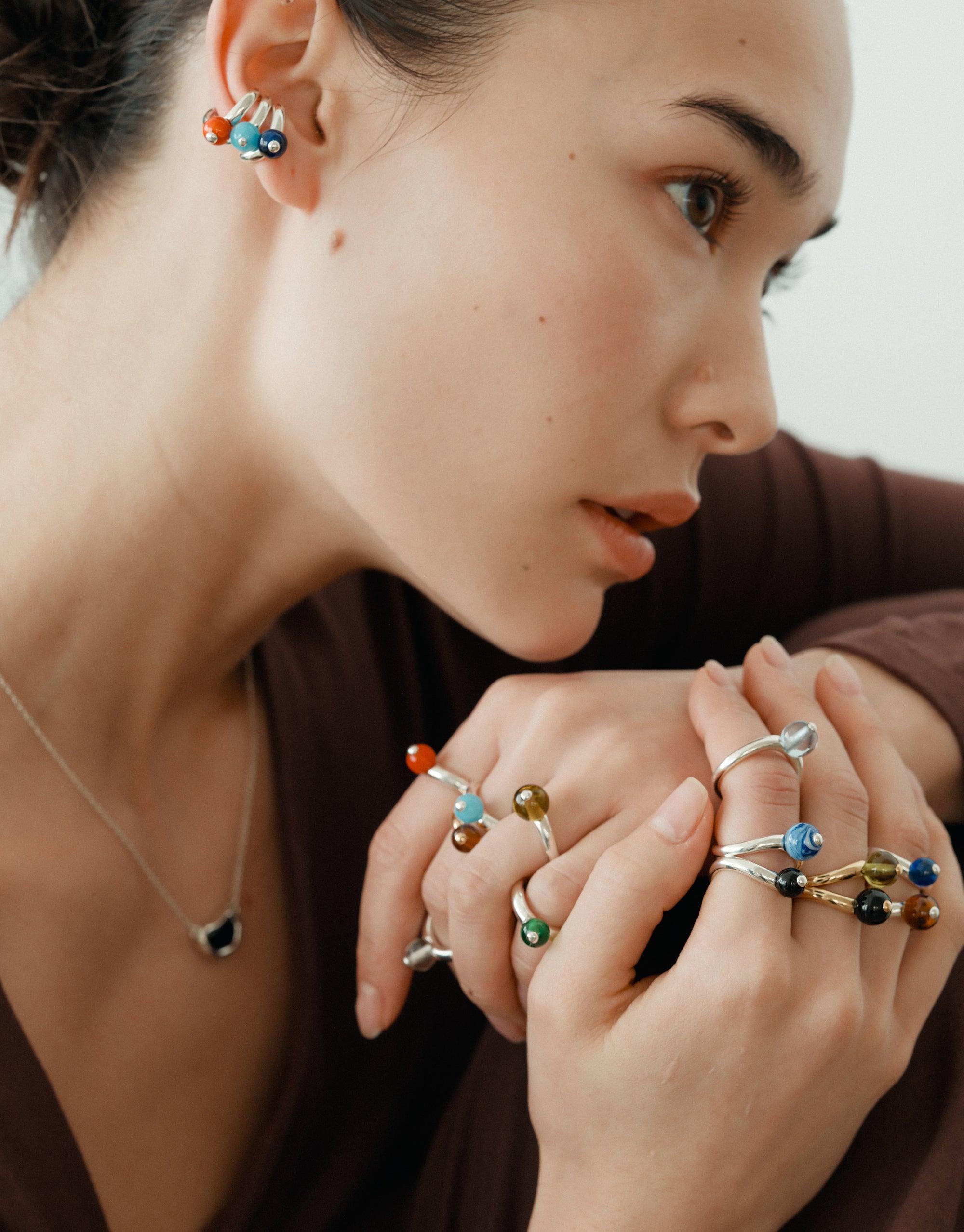 Close-up of a woman wearing multiple colorful rings and earrings inspired by organic shapes, showcasing the Bean Necklace collection. Her hands are positioned near her face, emphasizing the intricate designs and natural curves of the jewelry. CLED Bean Necklace