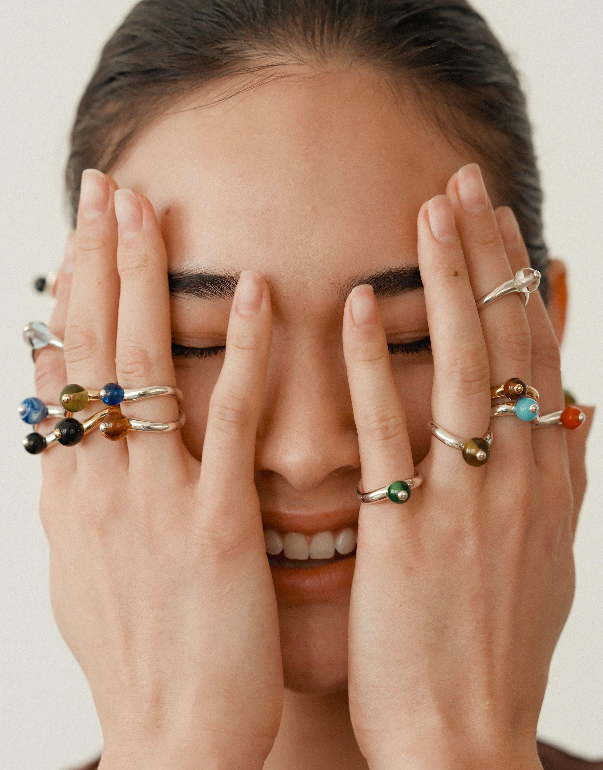 Close-up of hands adorned with multiple colorful rings, including the Pea Curve Ring | Large, covering eyes of a smiling person. Showcases unique jewelry designs inspired by nature's organic shapes and patterns. CLED