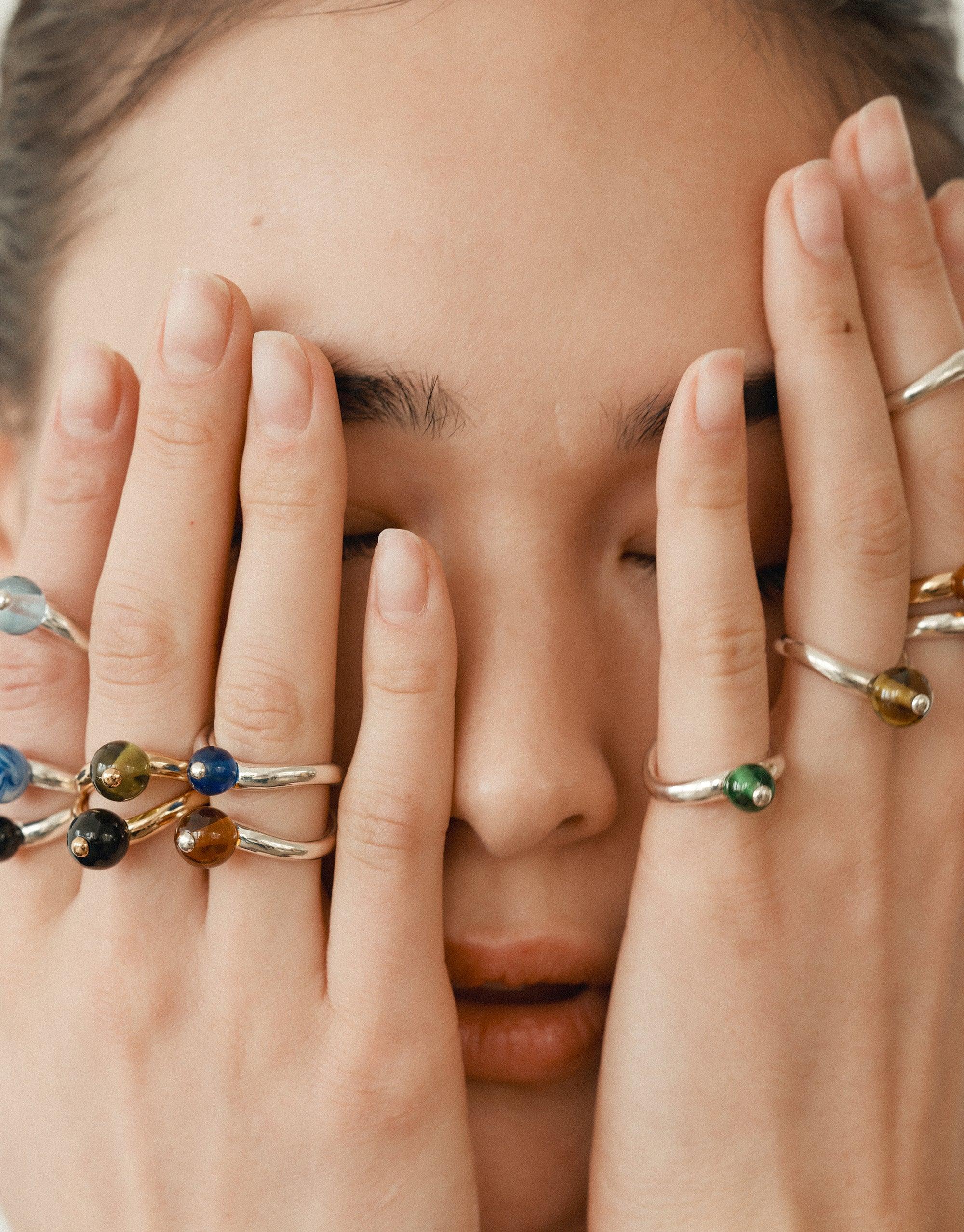 Close-up of hands with multiple colorful rings covering face, showcasing delicate curves and organic shapes inspired by nature's creativity. Pea Curve Ring Small featured among diverse ring designs, embodying growth and transformation. CLED