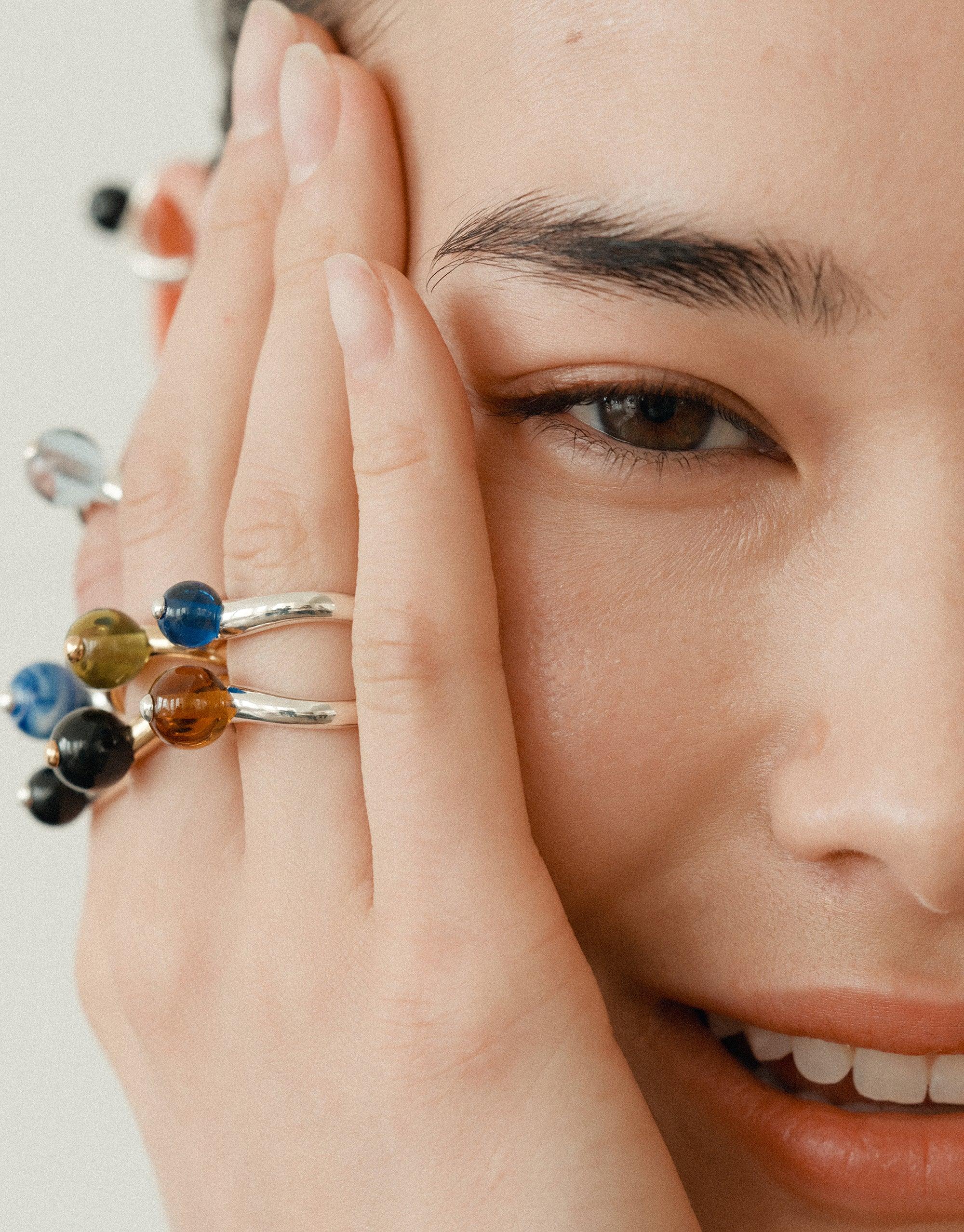 Close-up of a hand wearing multiple colorful rings, including the Pea Curve Ring | Small, showcasing the organic shapes inspired by nature's creativity. The rings feature curved designs and vibrant spherical elements, reflecting the collection's legume-inspired aesthetic. CLED