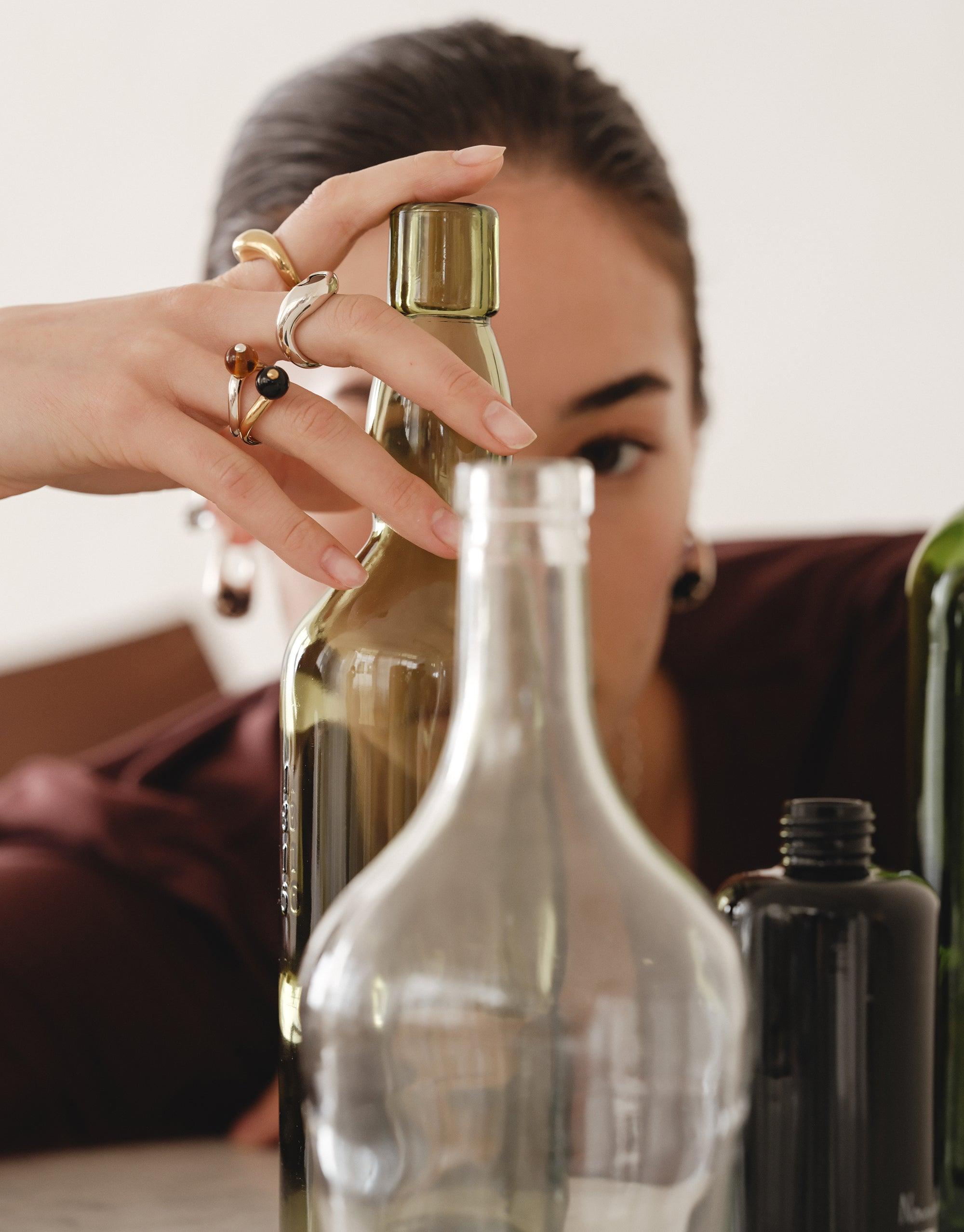 Hand adorned with elegant rings, including the Pea Curve Ring Large, reaching for a glass bottle. Showcases the organic, nature-inspired design against a backdrop of various bottles, capturing the essence of transformation and creativity. CLED