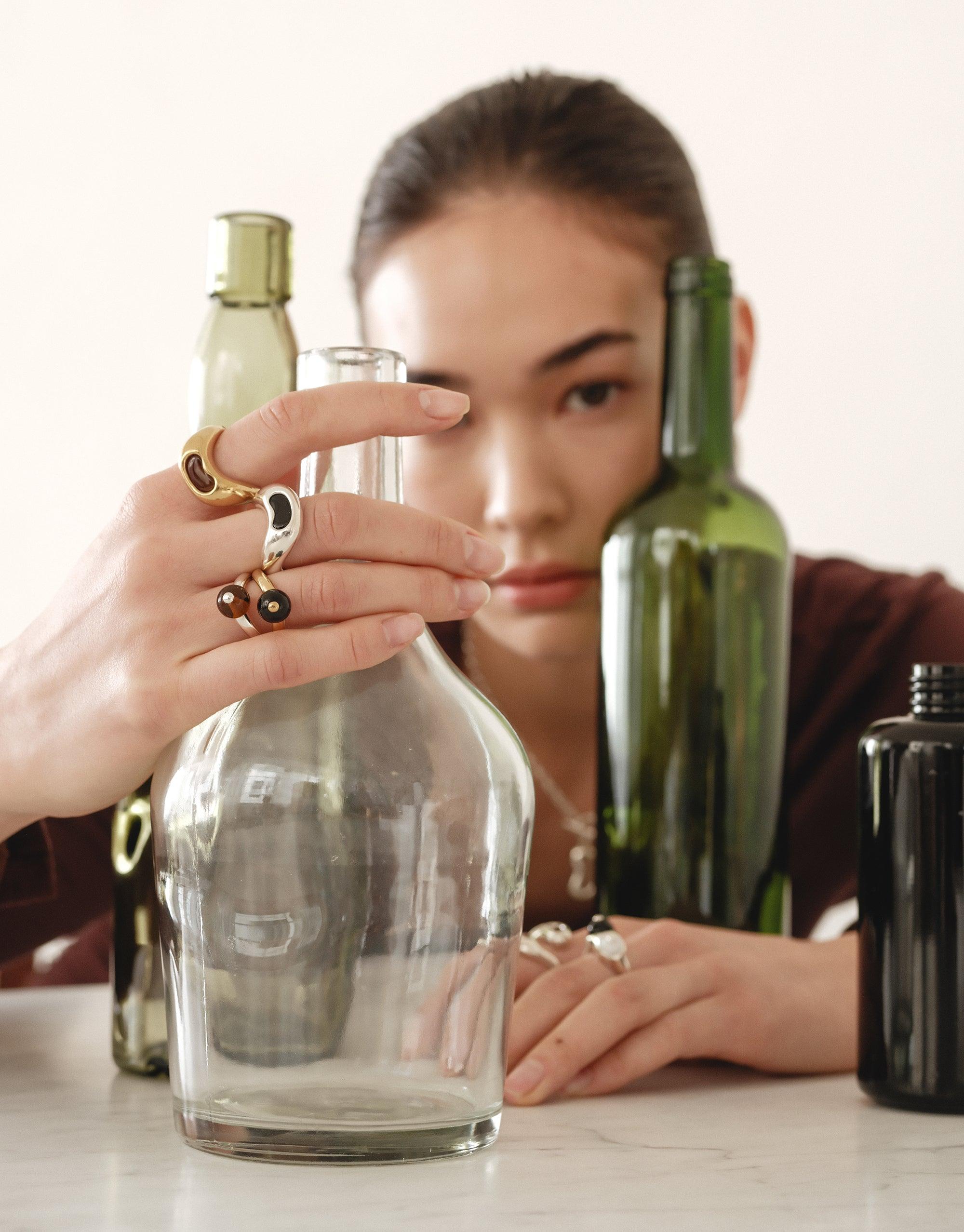 Person showcasing unique Bean Ring and other jewelry pieces, surrounded by glass and green bottles, demonstrating the natural-inspired design of the collection. Organic shapes reflect transformation and growth in legume-inspired jewelry. CLED Bean Ring