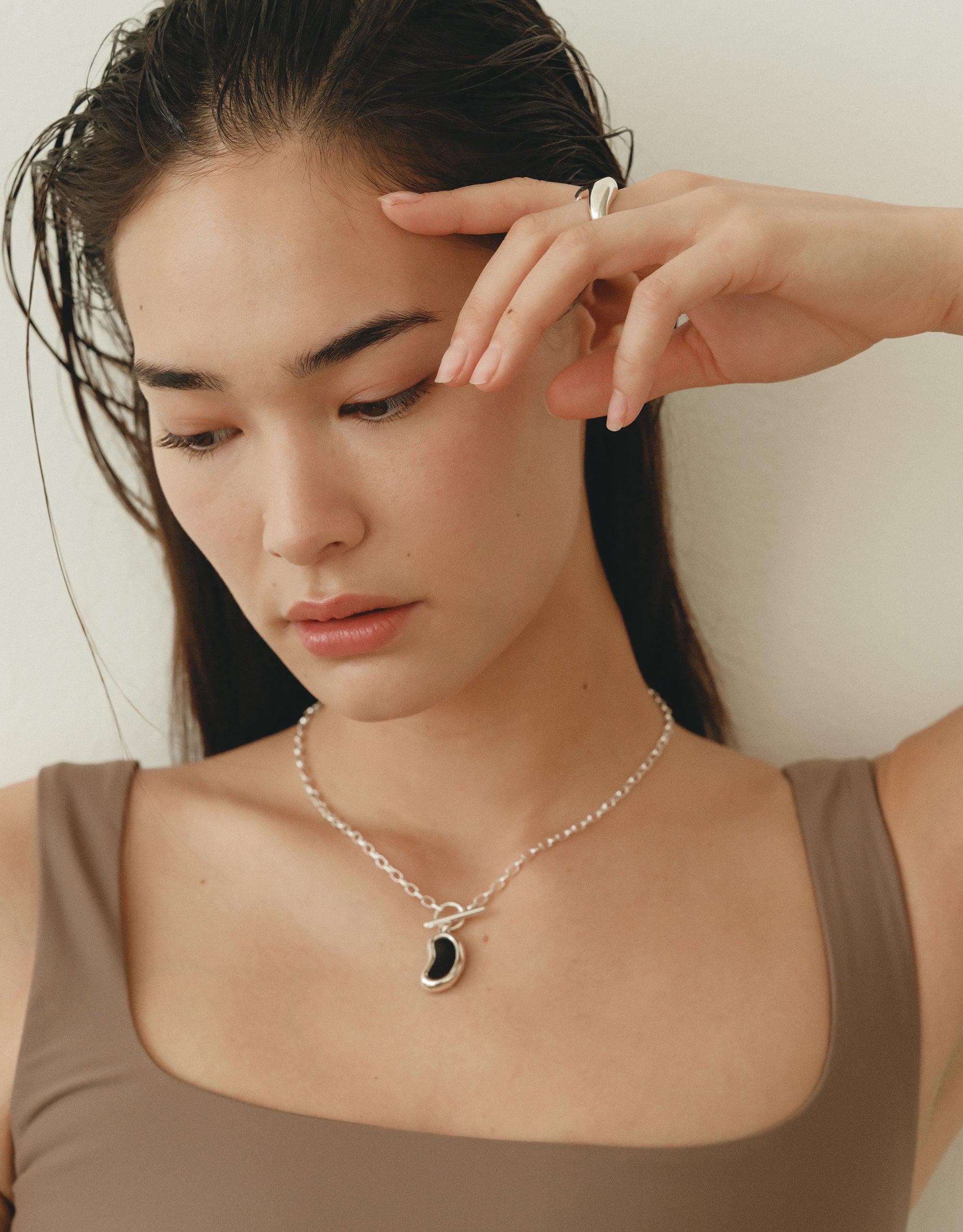 Elegant model wearing Bean Toggle Necklace, showcasing delicate silver chain with unique bean-shaped pendant. Natural, soft lighting highlights the necklace's organic design inspired by legume shapes. Perfect accessory for nature-inspired fashion. Bean Toggle Necklace by CLED.