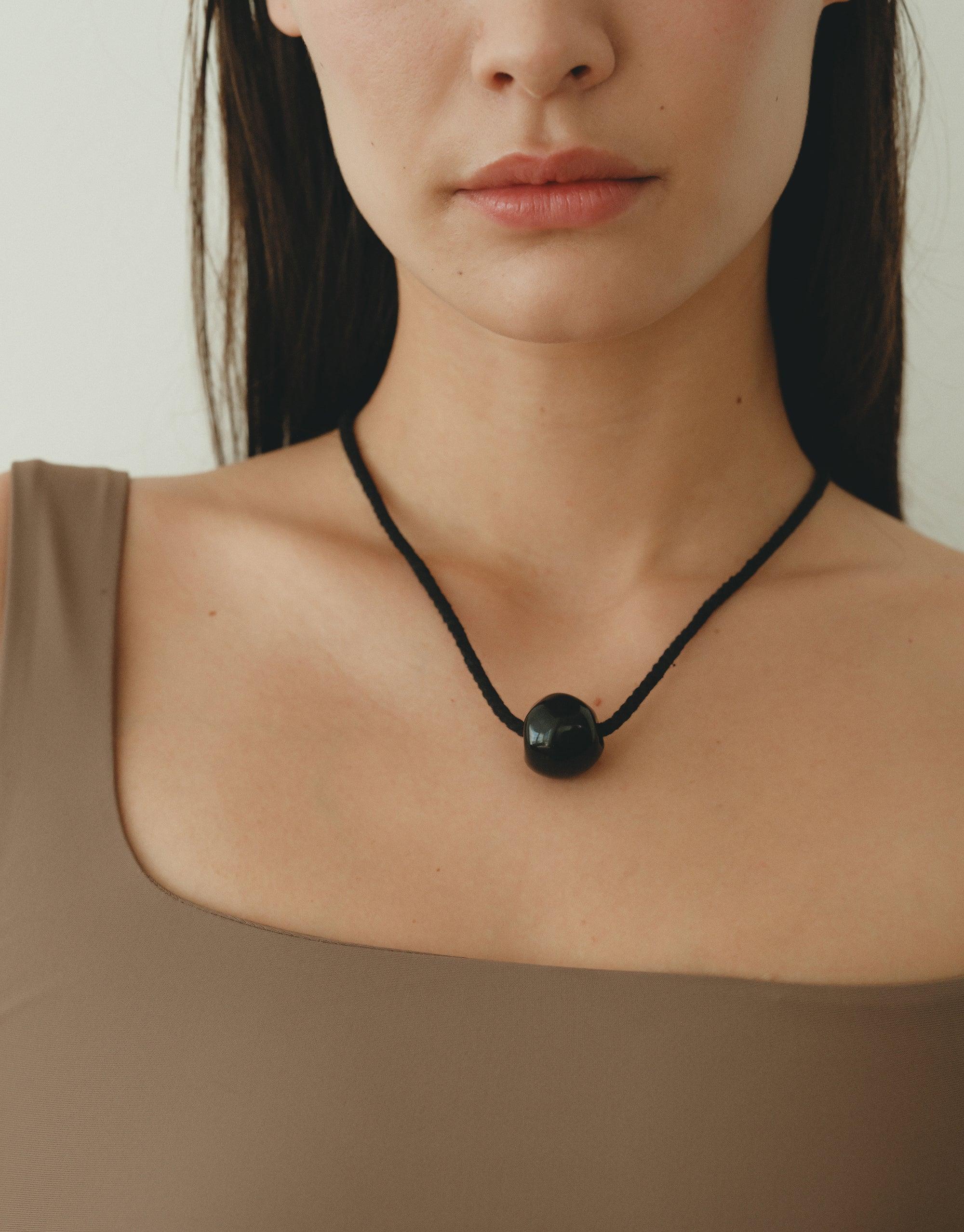 Elegant Black Pea Necklace featuring a single spherical pendant on a dark cord, inspired by organic legume shapes. Minimalist design perfectly complements neutral attire, embodying natural beauty and transformation. Black Pea Necklace by CLED.
