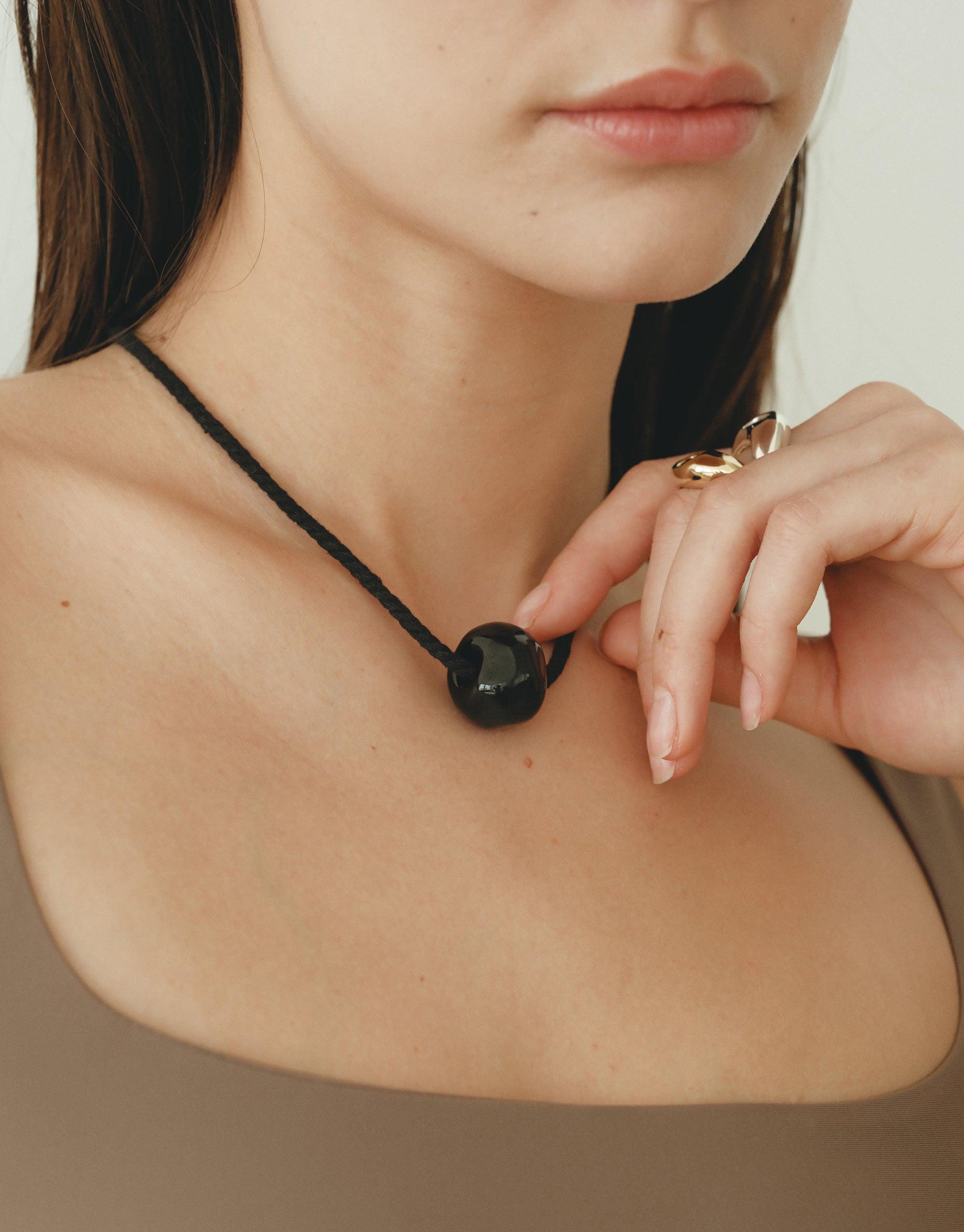 Close-up of a person wearing the Black Pea Necklace, showcasing its organic shape inspired by nature's creativity. The necklace features a dark, spherical pendant on a thin black cord, elegantly resting against the wearer's collarbone. Black Pea Necklace by CLED.