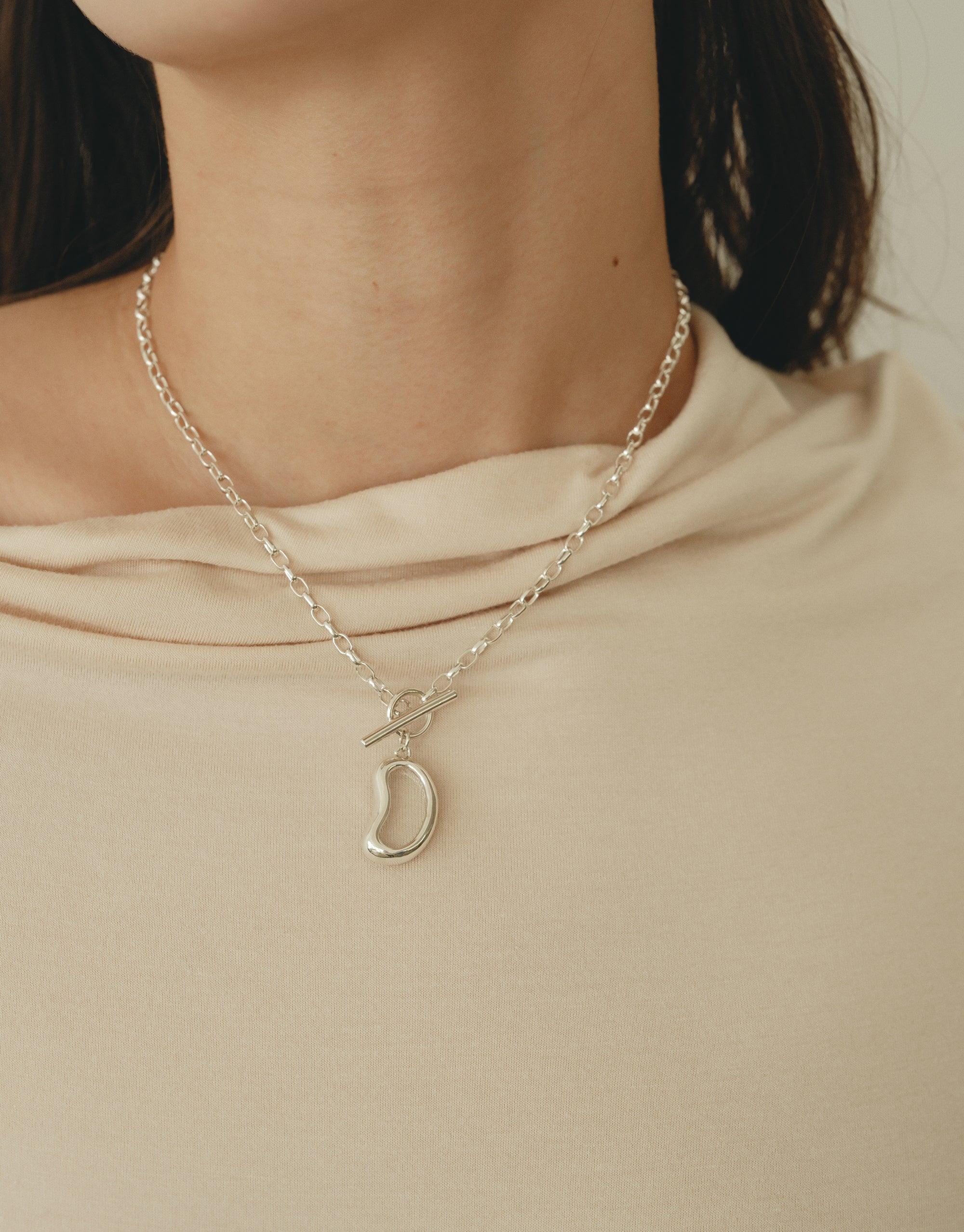 Elegant silver Bean Toggle Necklace with delicate chain and organic bean-shaped pendant, showcasing nature-inspired design. Worn against beige fabric, the necklace embodies transformation and growth. Perfect for nature lovers and minimalist jewelry enthusiasts. Bean Toggle Necklace by CLED.
