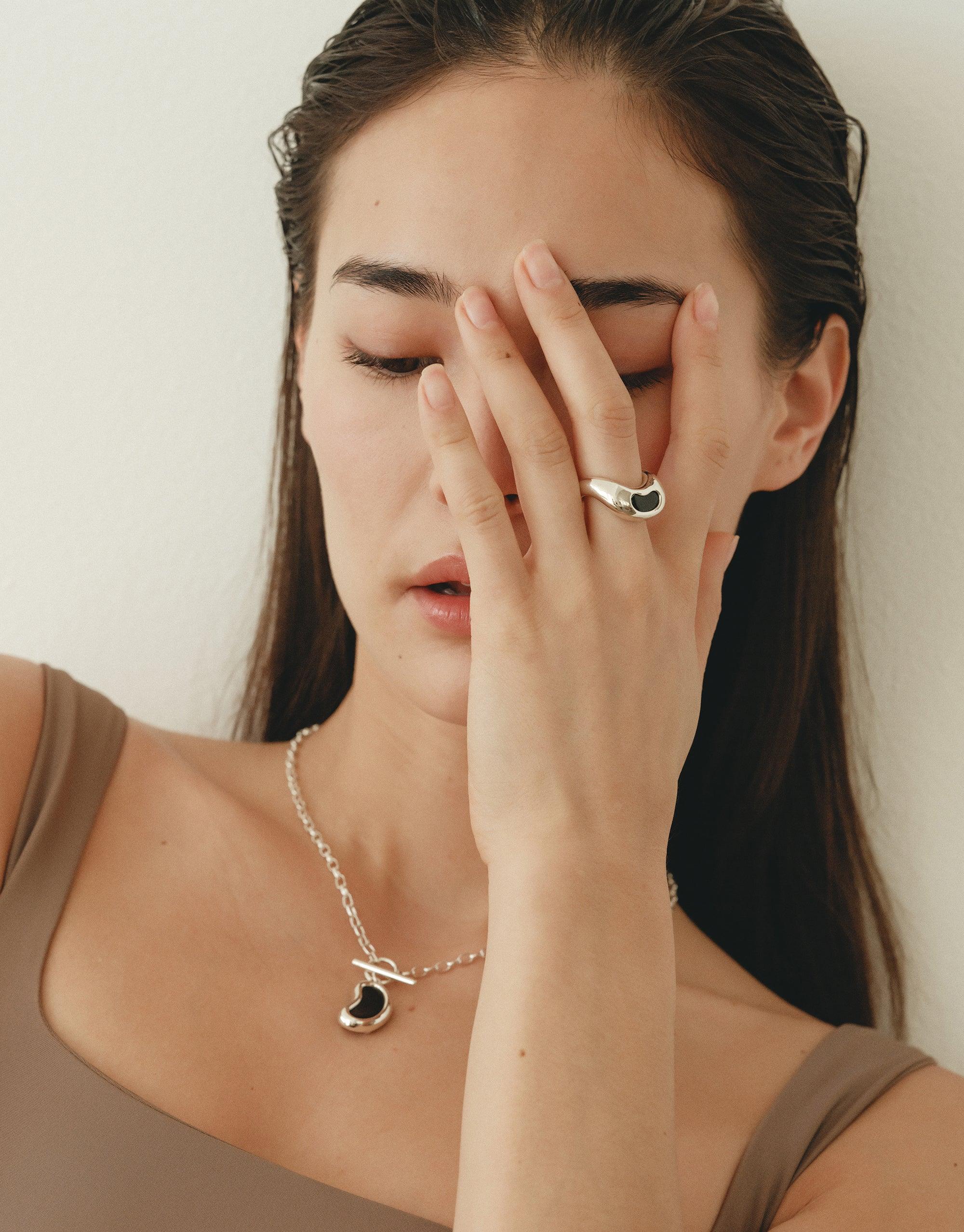 Woman wearing Bean Ring, showcasing unique organic shape inspired by legume plants. Elegant silver jewelry design on hand, complemented by matching necklace. Natural beauty and transformation embodied in stylish accessory. Bean Ring by CLED