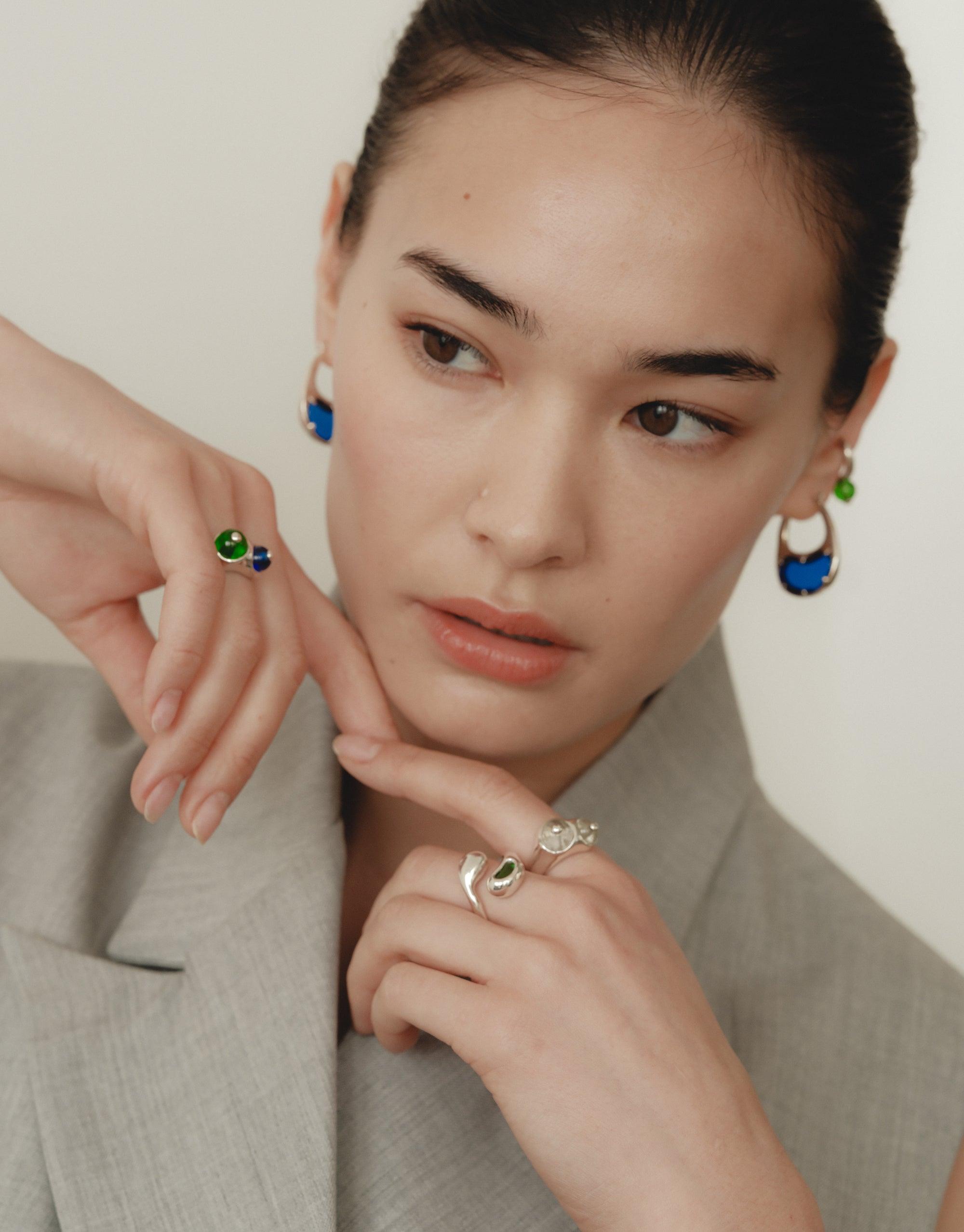 Sprout Ring on a woman's hand, showcasing its elegant design inspired by legume shapes, emphasizing nature's transformative beauty, part of CLED's eco-friendly jewelry collection made from repurposed glass. Women is also wearing blue tone hoop earrings