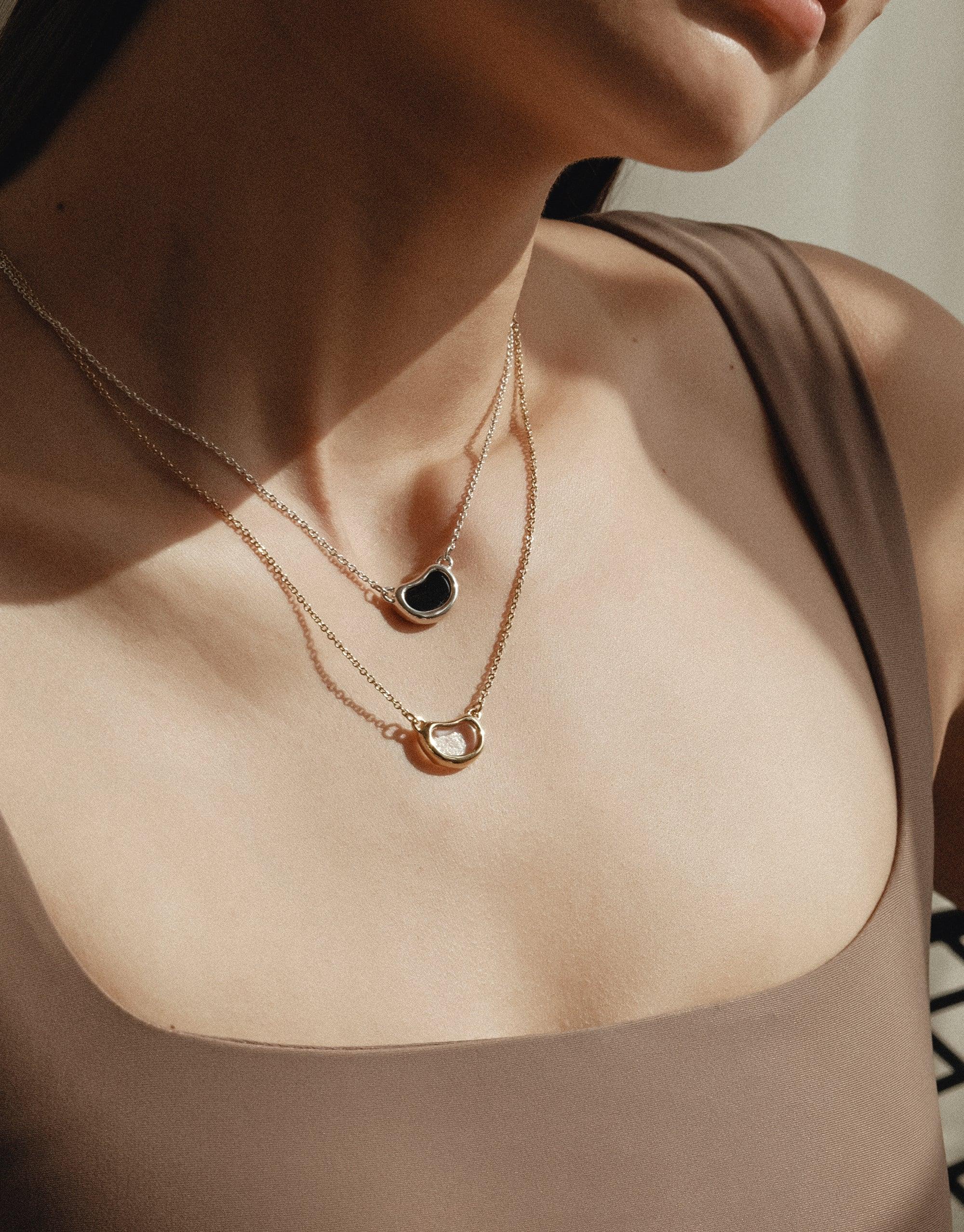 Close-up of a person wearing layered delicate gold necklaces, featuring organic bean-shaped pendants in contrasting colors. The necklaces rest elegantly on the collarbone, showcasing the Bean Necklace's design inspired by nature's soft curves and patterns. CLED