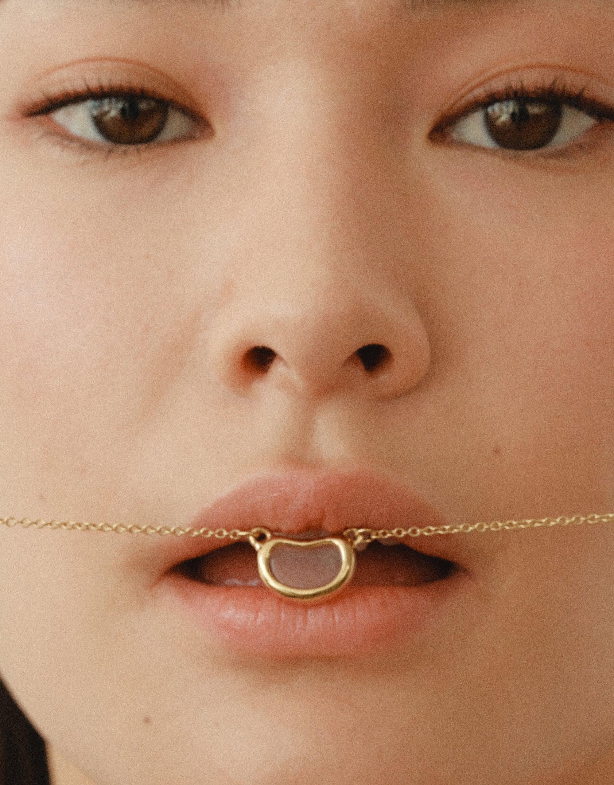 Close-up of lips wearing a delicate gold Bean Necklace, showcasing its organic shape inspired by nature's creativity. The necklace rests gently on parted lips, highlighting its elegant design and connection to natural forms. CLED Bean Necklace.