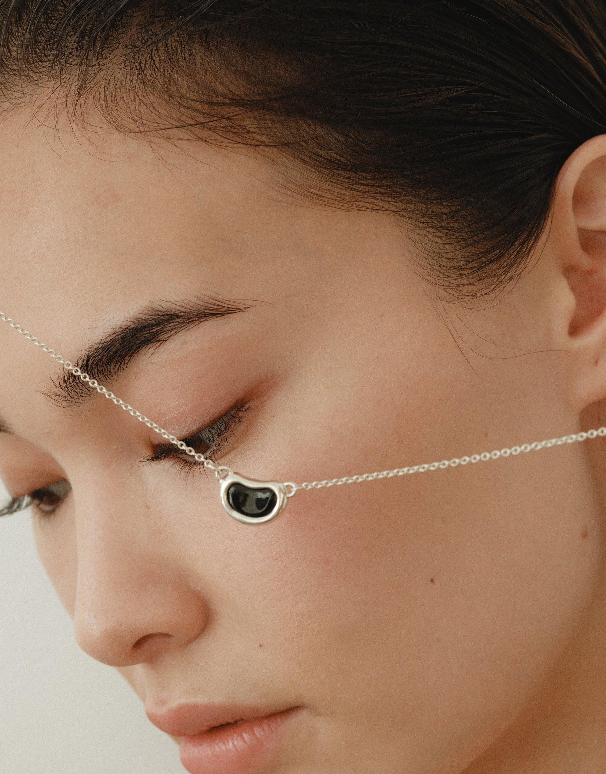 Close-up of a delicate silver chain necklace with a small bean-shaped pendant, showcasing the Bean Necklace's elegant design inspired by nature's organic forms. The necklace rests gently across the face, highlighting its minimalist beauty and versatile wearability. CLED