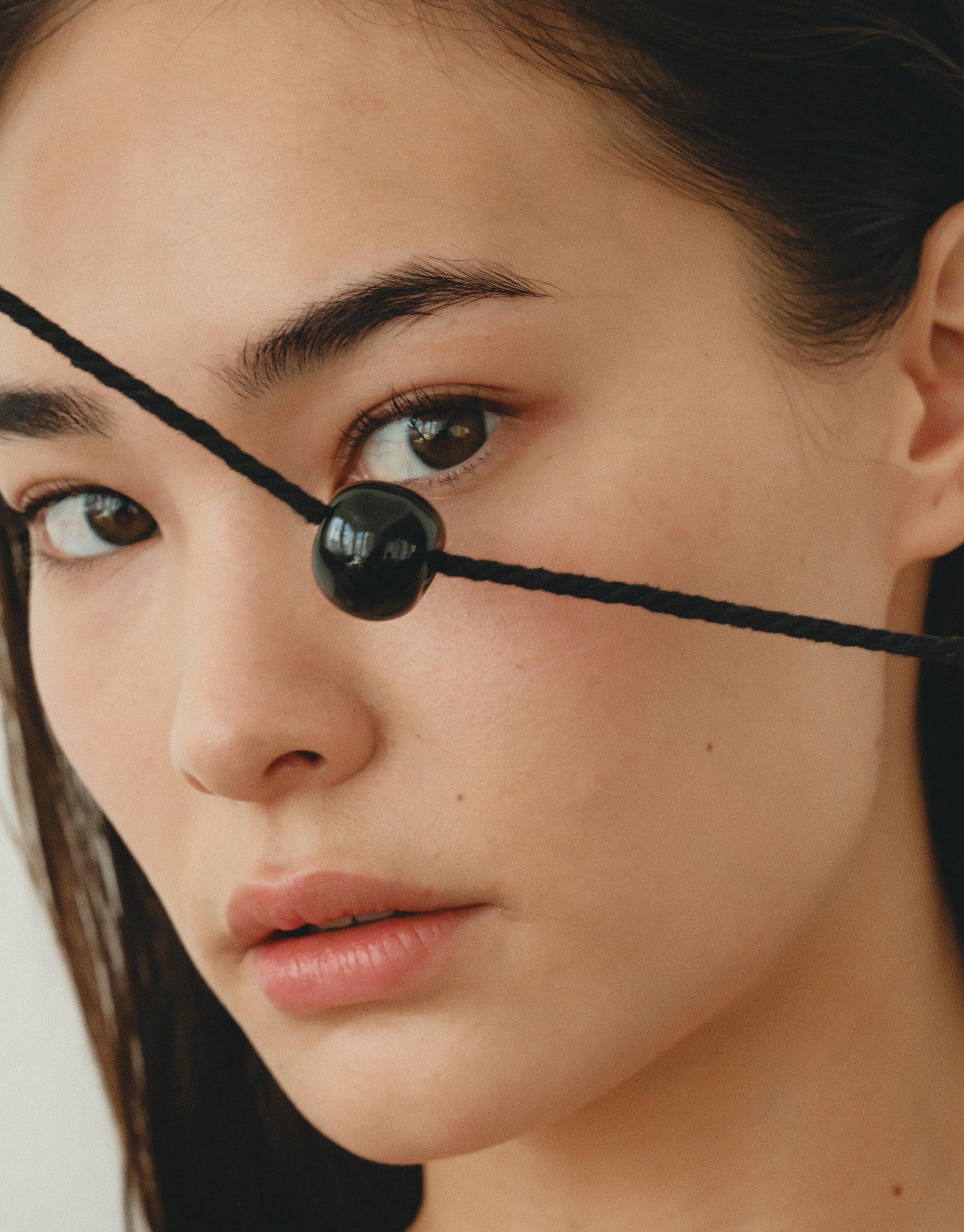 Close-up of a model wearing the Black Pea Necklace, showcasing a sleek black bead on a thin cord across the face, highlighting the necklace's unique and minimalist design inspired by nature's organic shapes. Black Pea Necklace by CLED.