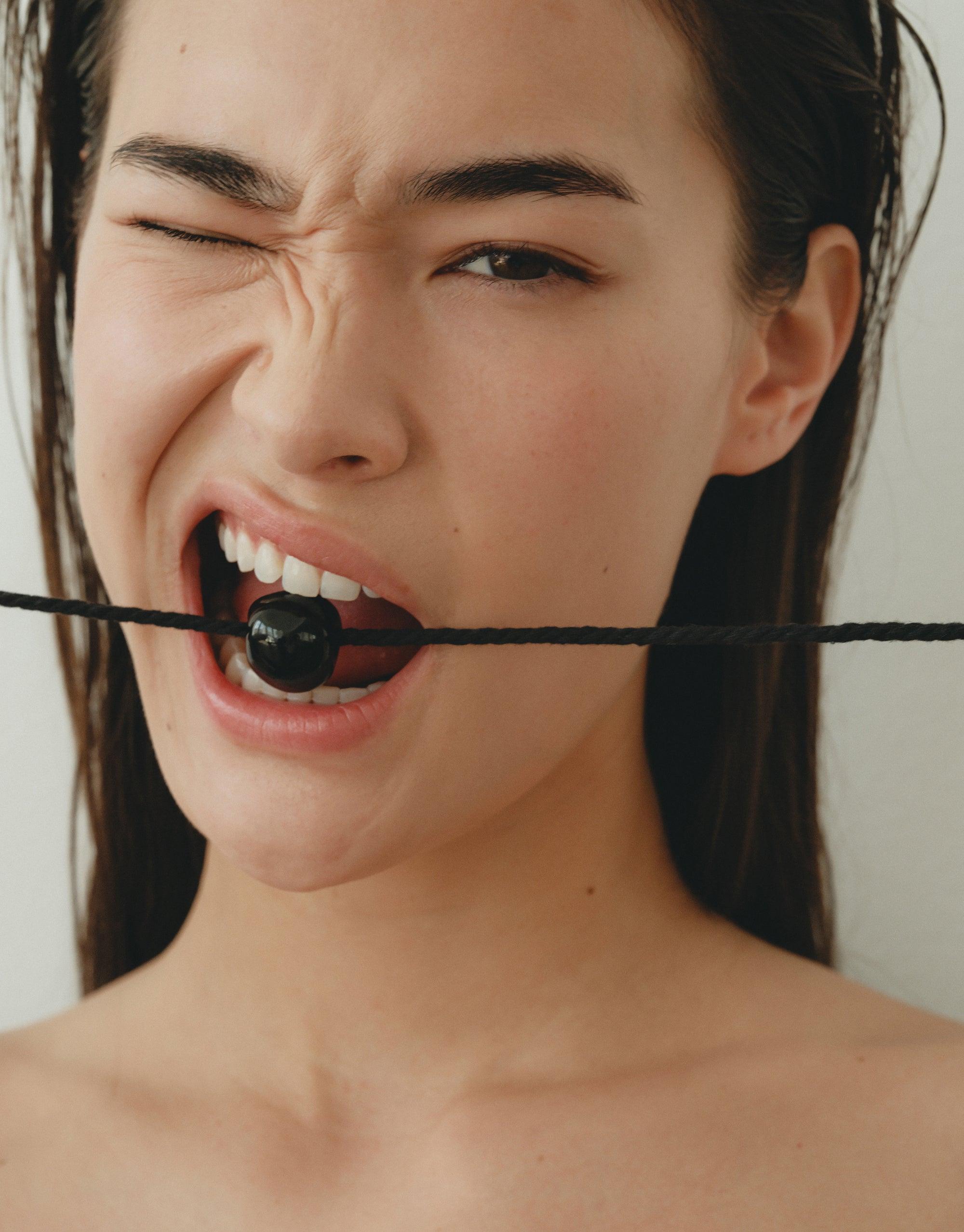 Close-up of a person biting a black spherical object suspended on a thin cord, showcasing the unique design and wearability of the Black Pea Necklace. The image captures the organic shape and natural inspiration behind the legume-inspired jewelry collection by CLED.