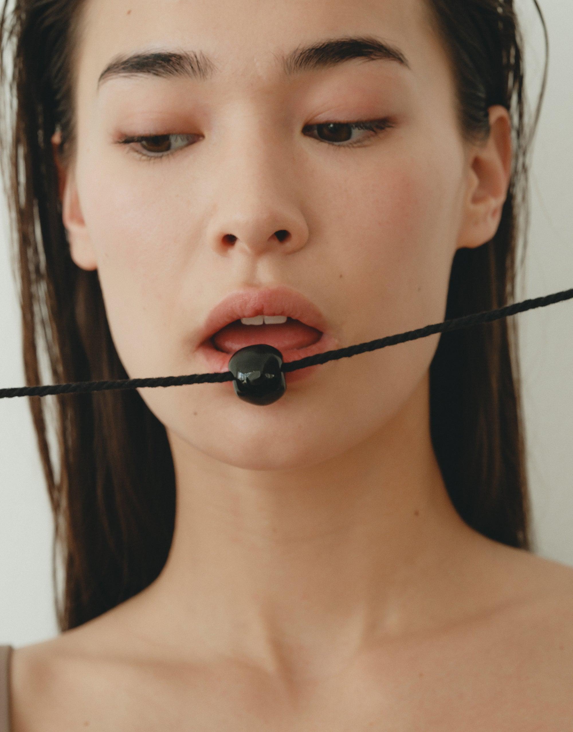 Close-up of a model wearing the Black Pea Necklace, showcasing the unique legume-inspired design on a black cord. The necklace rests gently between the lips, highlighting its organic shape and natural aesthetic. Black Pea Necklace by CLED.