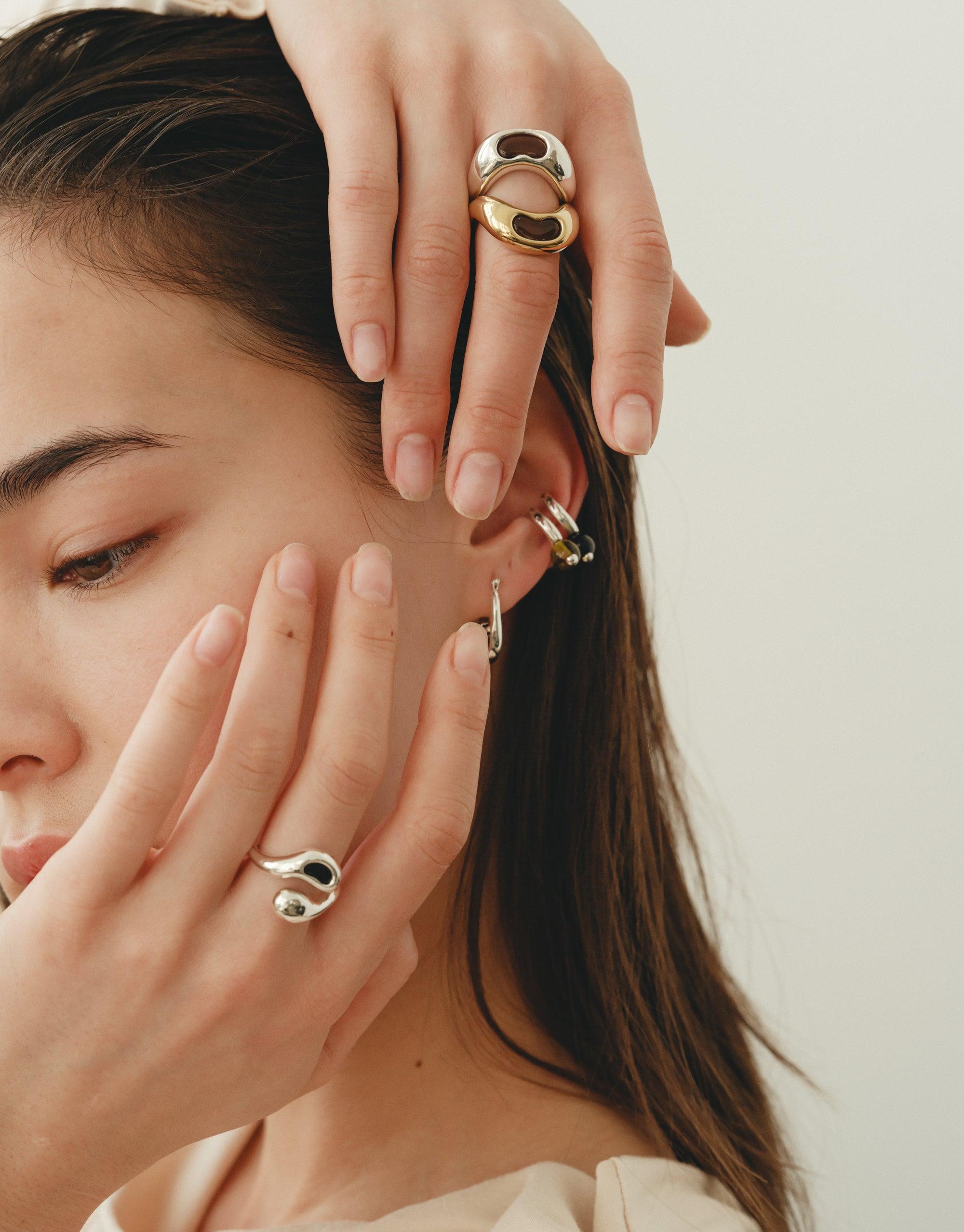 Close-up of hands adorned with elegant Bean Rings, showcasing organic shapes inspired by nature. Earrings complement the rings, highlighting the collection's versatile design. Natural beauty and creativity reflected in jewelry. Bean Ring by CLED.