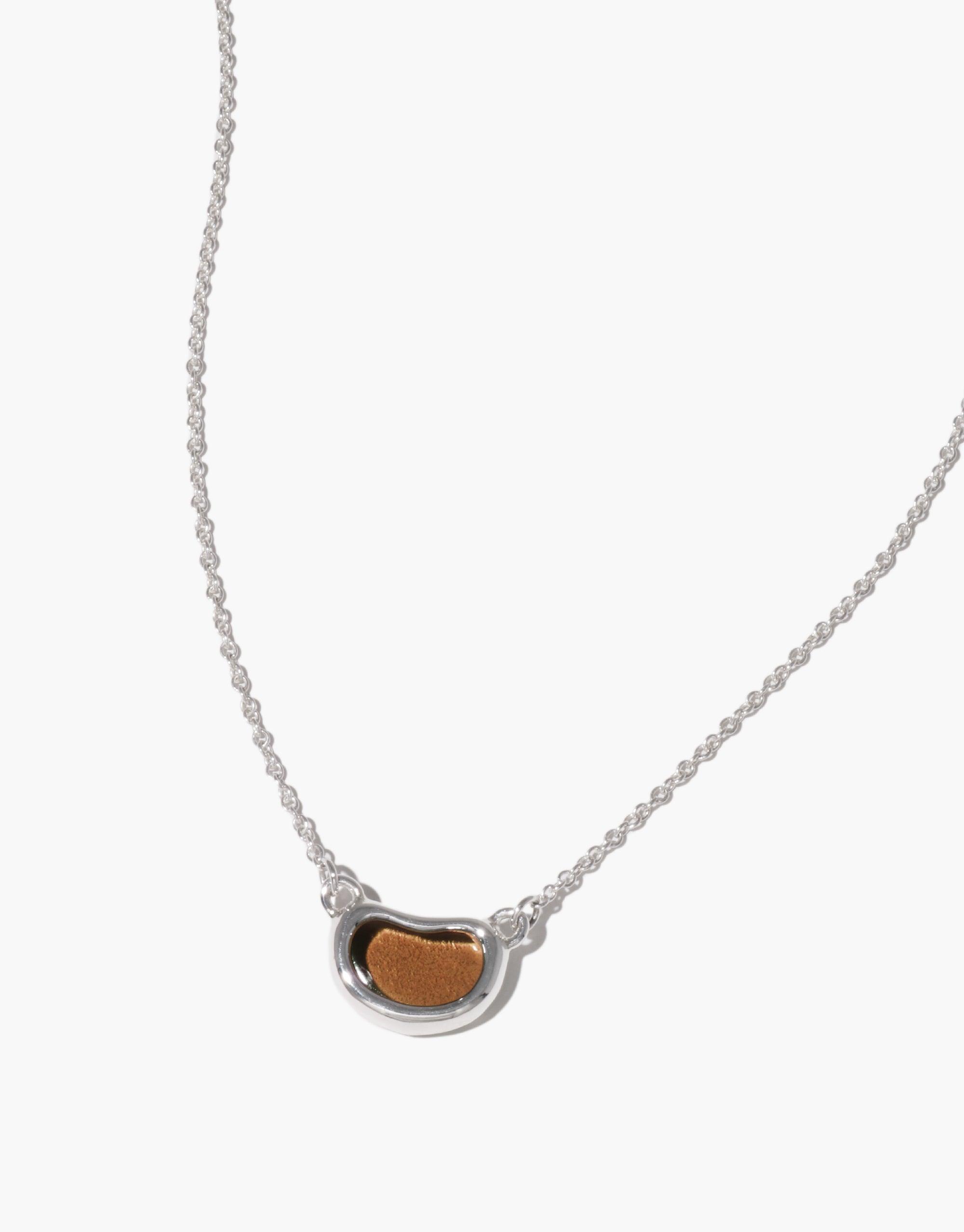 Elegant Bean Necklace featuring a delicate silver chain with a small, bean-shaped pendant in silver and warm brown tones, inspired by nature's organic forms and growth patterns. A unique piece of jewelry that captures the essence of transformation and natural beauty. CLED Bean Necklace.