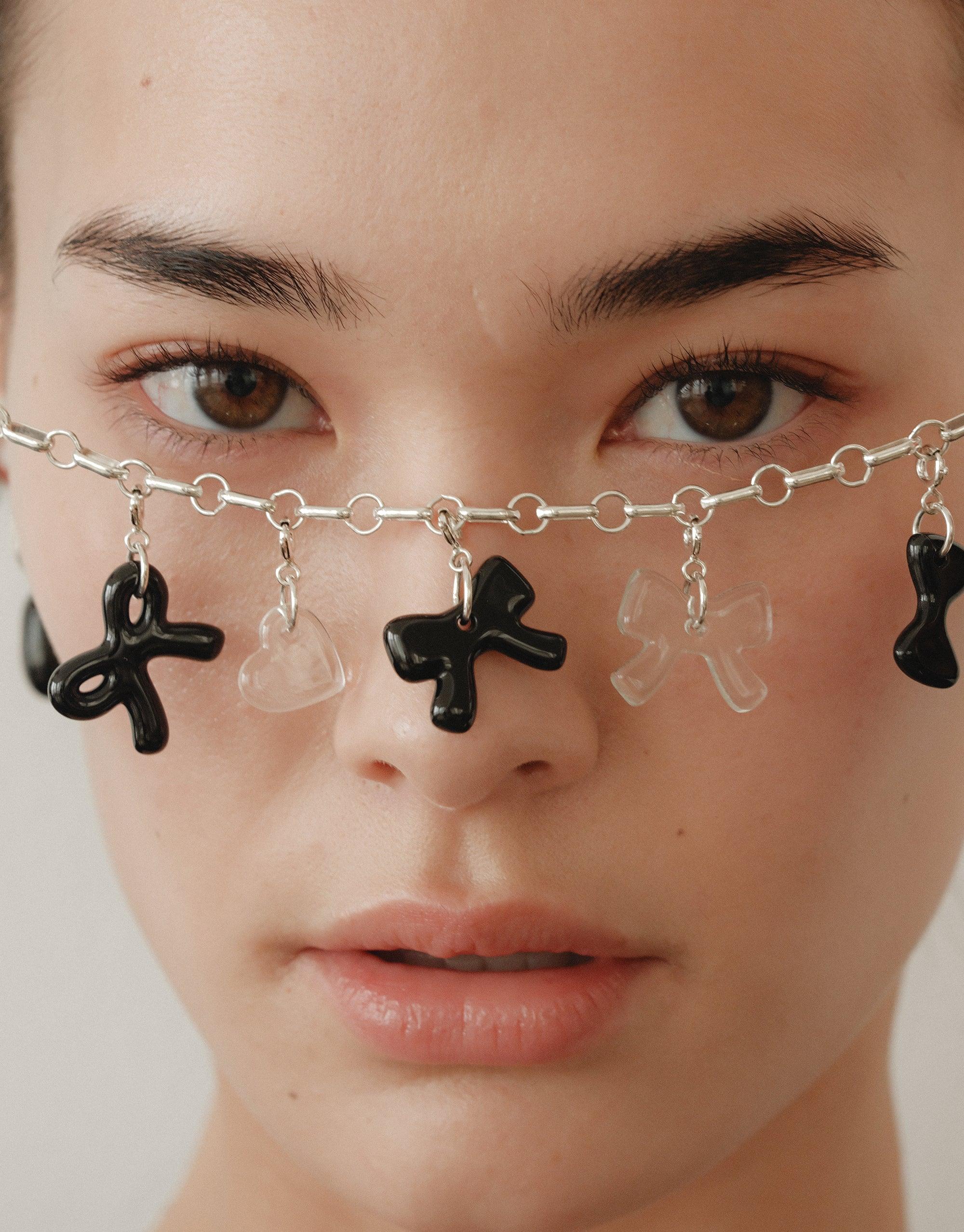 Close-up of a silver chain necklace with black and clear bow-shaped charms, displayed across the bridge of a person's nose. Showcases the playful and distinctive Bow A Icon Charm design for enhancing jewelry. CLED