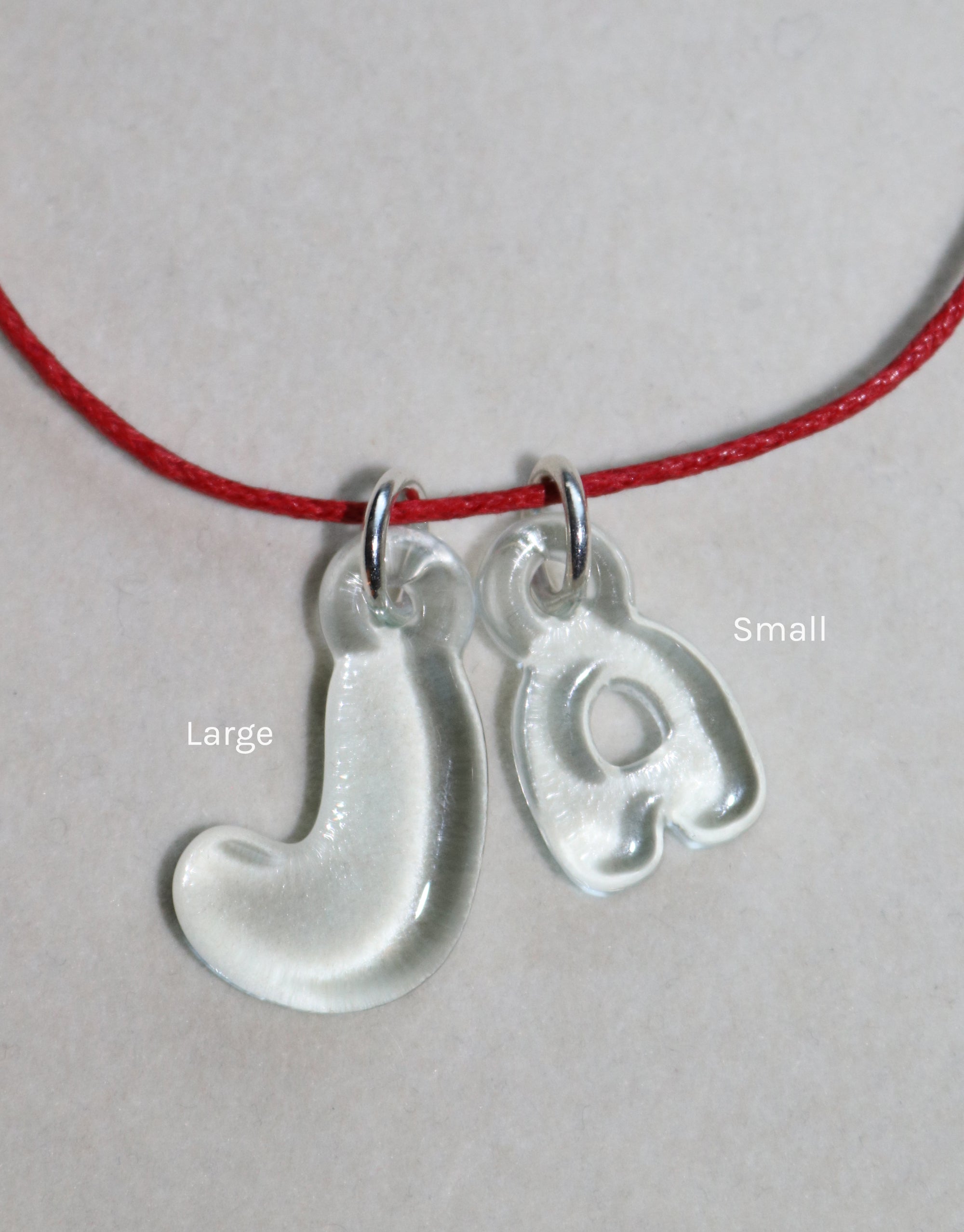 Eco-friendly bubble letter charms 'J' and 'a' in large and small sizes, made from recycled window glass, hanging on a red cord necklace. Playful and personalized jewelry for everyday wear. Letter Charm | Alphabet by CLED