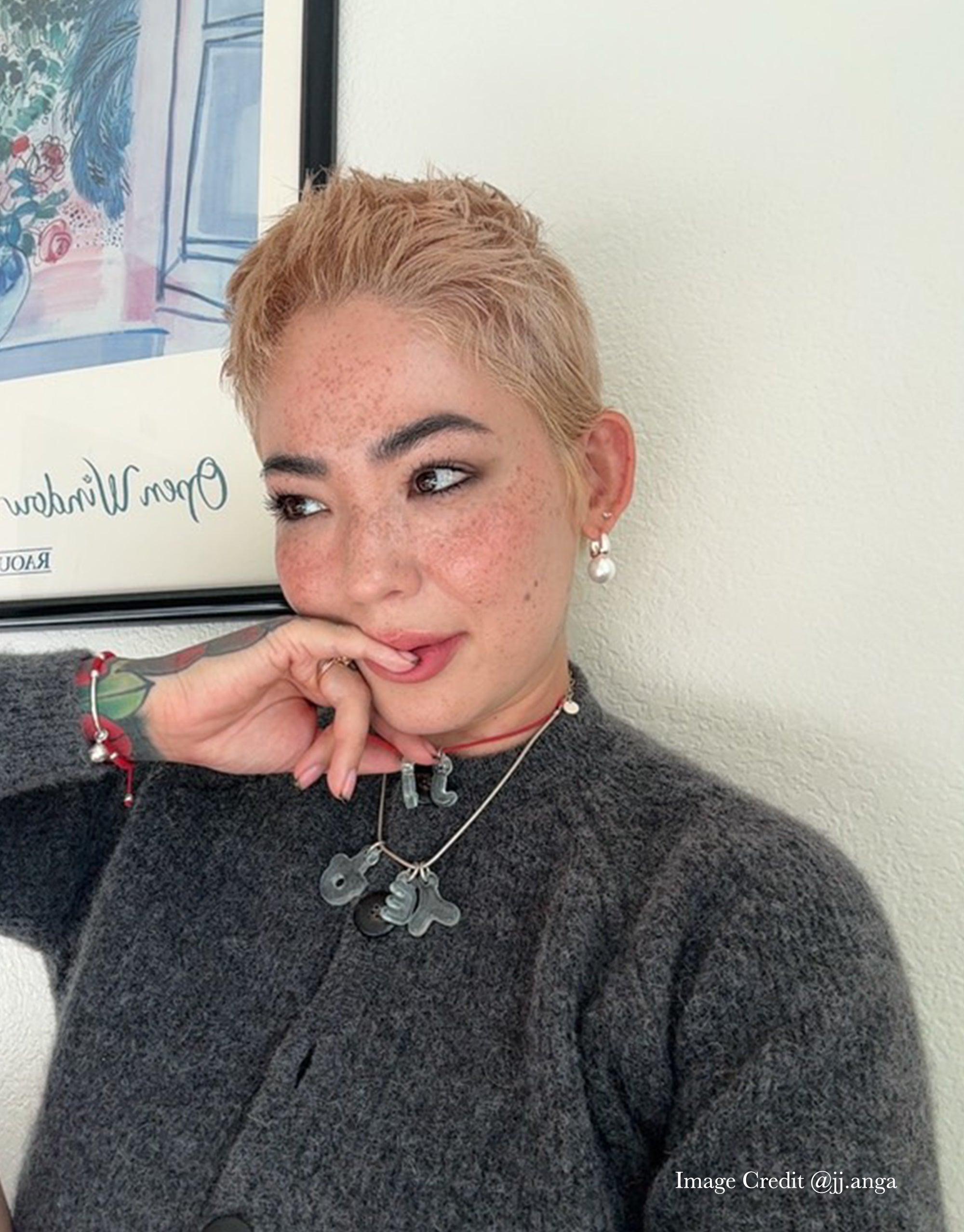 Close-up portrait showcasing Letter Charm | 한글 Hangul necklace with multiple glass charms. Model wearing gray sweater, short blonde hair, and freckles. Earrings and colorful bracelet visible. Background includes wall art. Eco-friendly jewelry celebrating Korean culture and personal expression. CLED