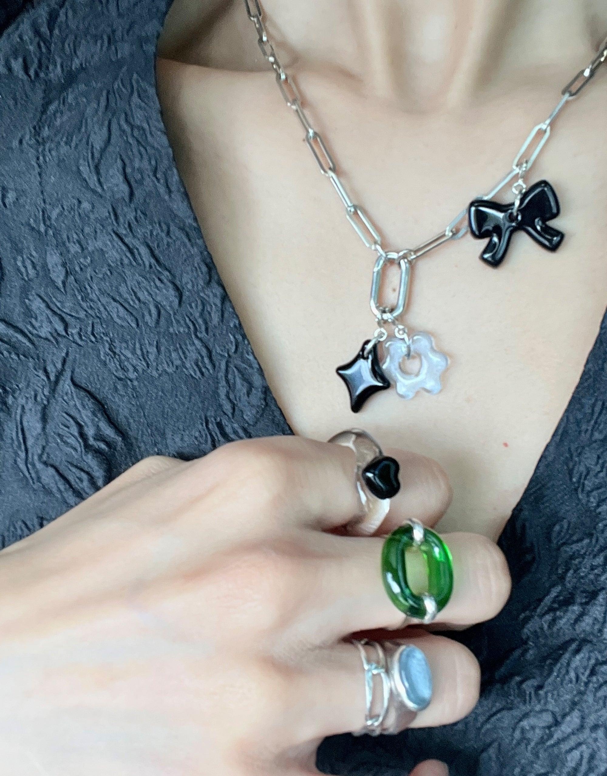 Stylish silver chain necklace with black bow, star, and flower charms, worn with colorful rings. Versatile Icon Chain Necklace for customizable style. Create a bold statement with this CLED Icon Chain Necklace | D.