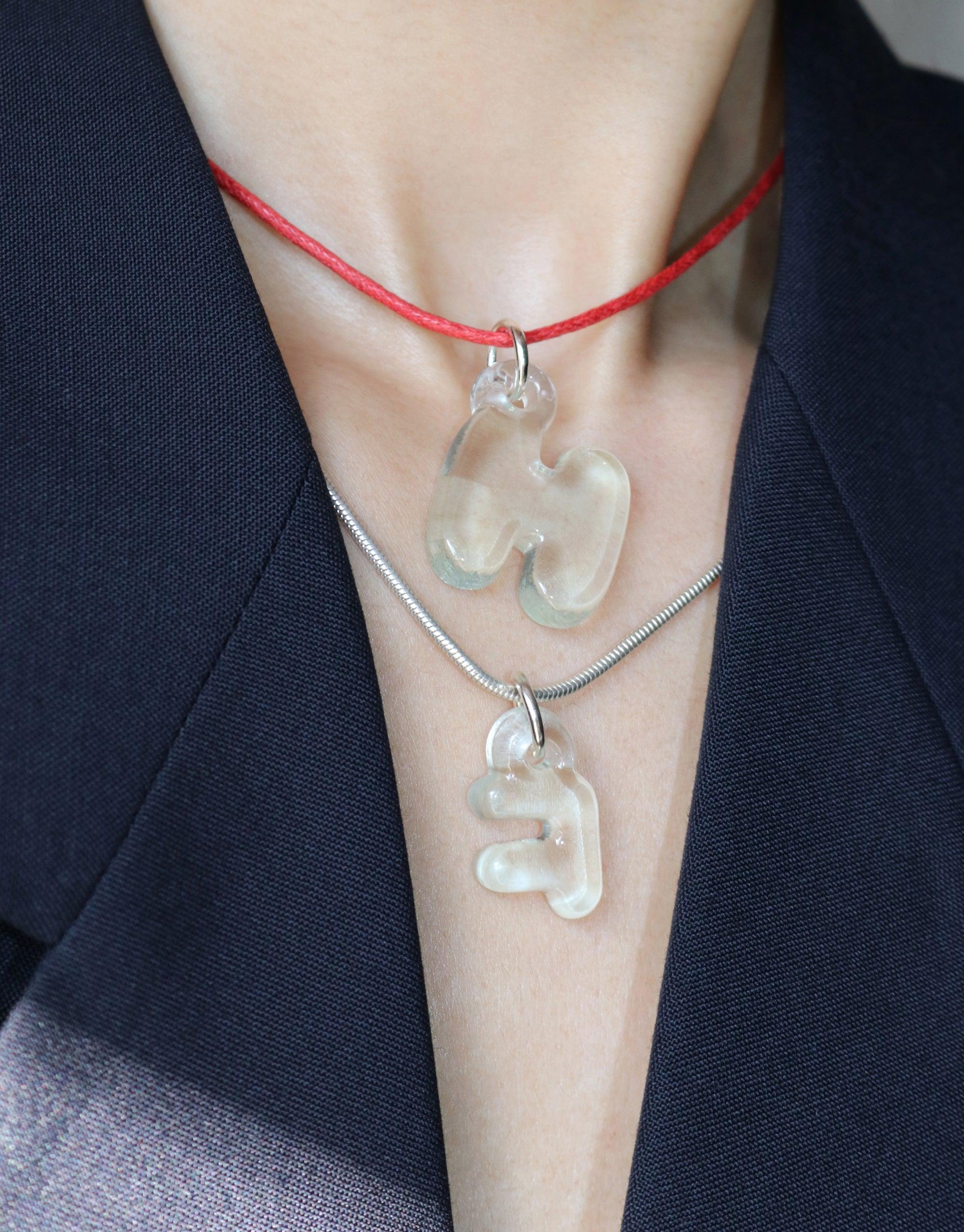 Adjustable cord necklace with transparent letter charms displayed on model, showcasing versatile layering options. Red cotton cord and silver snake chain demonstrate length variety. Perfect for customizable charm accessories. Cord Necklace by CLED.