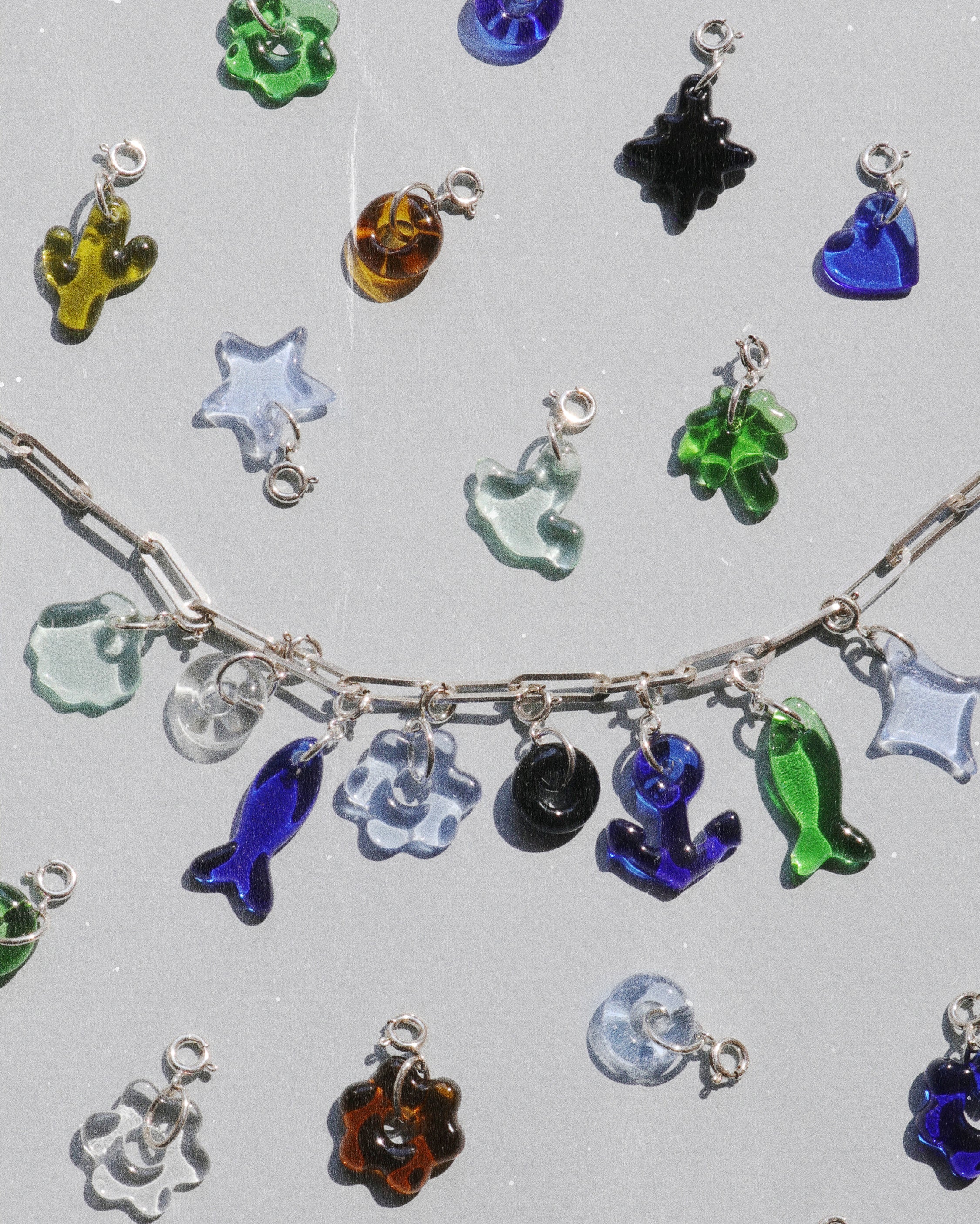 Handcrafted upcycled glass charms in blue, green, clear, amber, and black tones displayed with a silver chain, featuring playful shapes like fish, stars, hearts, and organic forms.