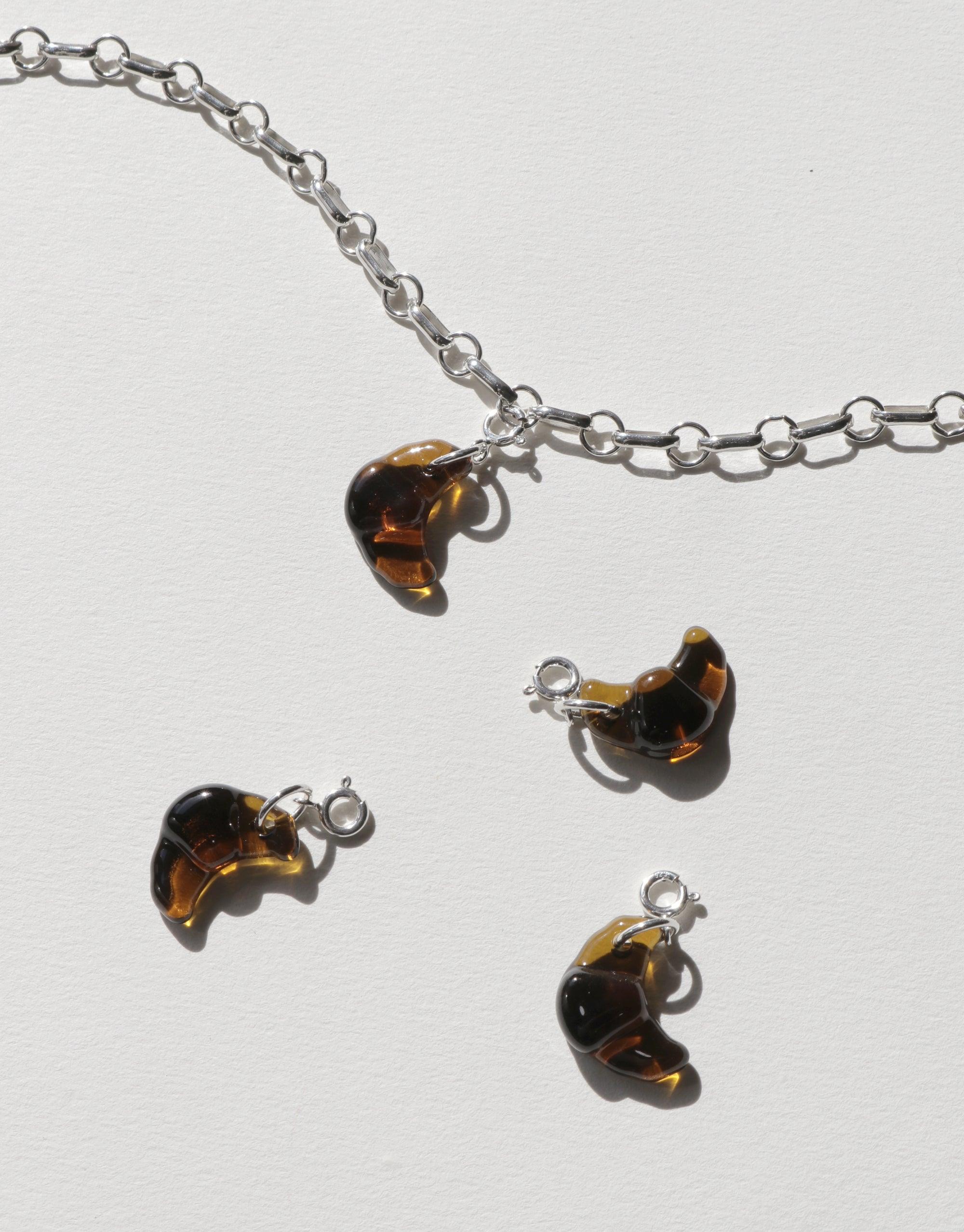 Silver chain necklace with amber-colored croissant-shaped charm, accompanied by three additional croissant charms. Playful and quirky Icon Charm design for versatile jewelry customization. Croissant | Icon Charm by CLED.