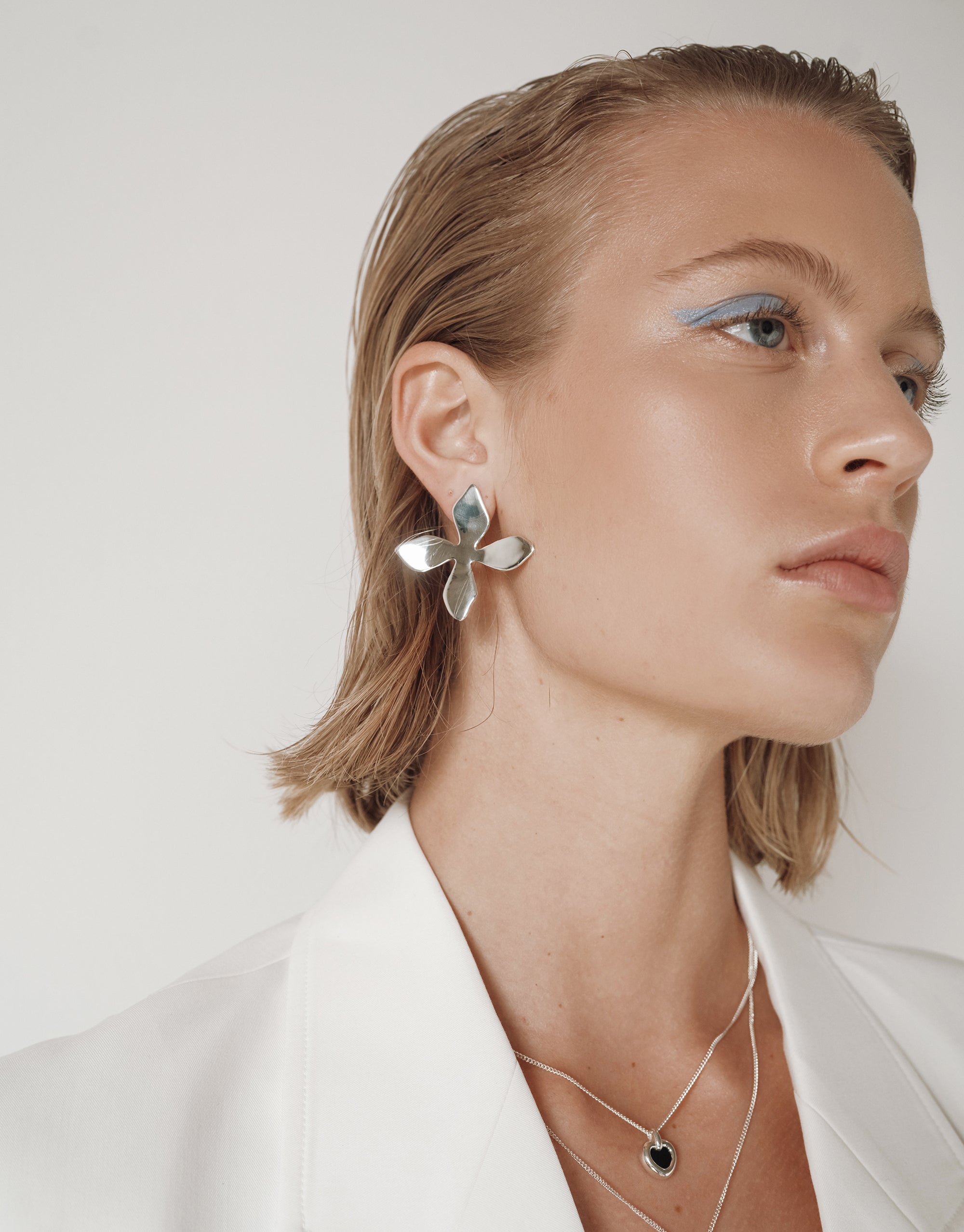 Close-up of a woman wearing bold, reflective silver flower-shaped earrings and layered silver necklaces, including a heart-shaped black pendant, styled with a sleek white blazer and minimal blue eyeliner.
