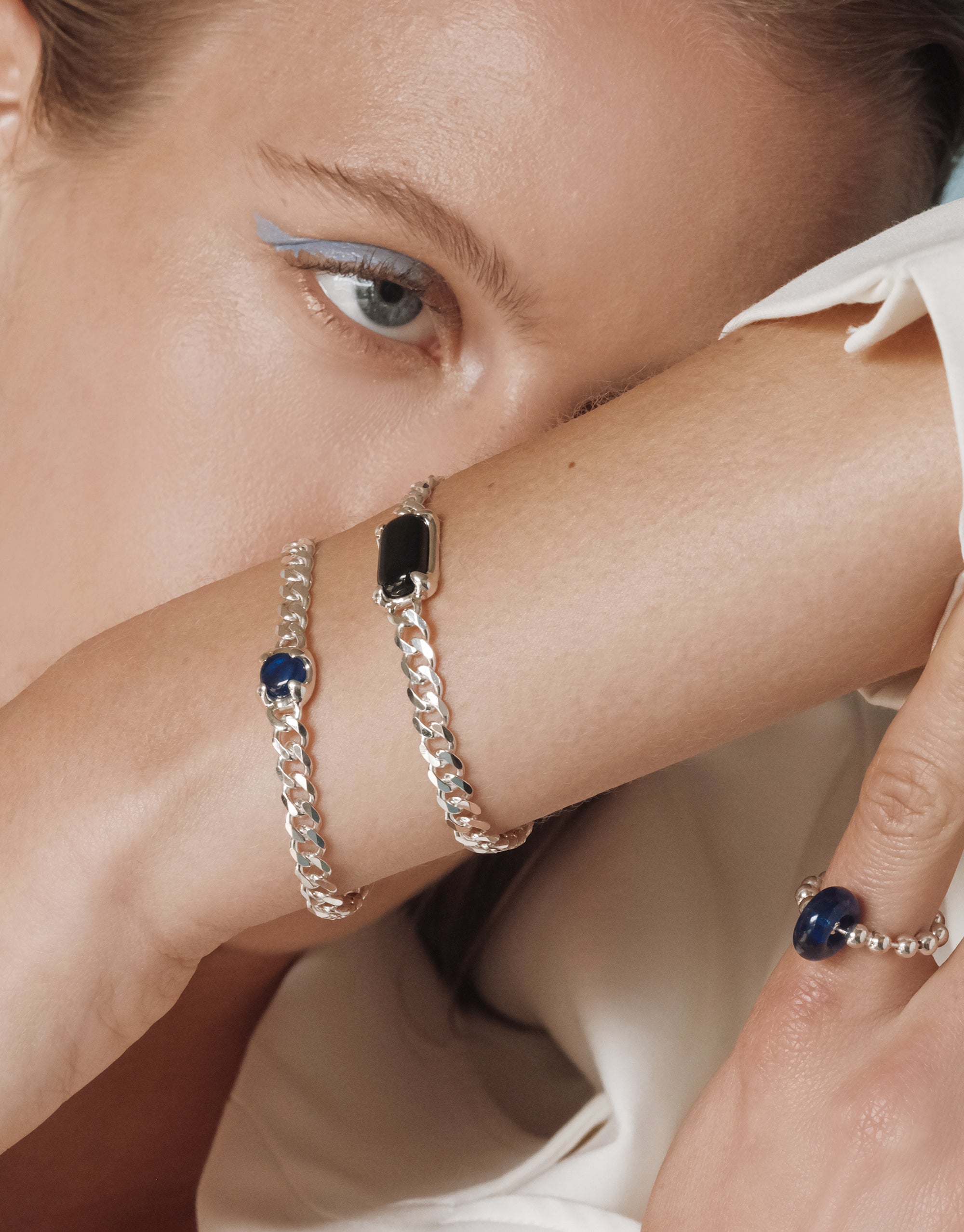 Close up of a model’s wrist and hand adorned with two silver chain bracelets, one with a deep blue Eco Gem and one with a black rectangular gem, paired with a beaded silver ring featuring a translucent blue glass bead. Styled with a white outfit and pastel blue eyeliner.