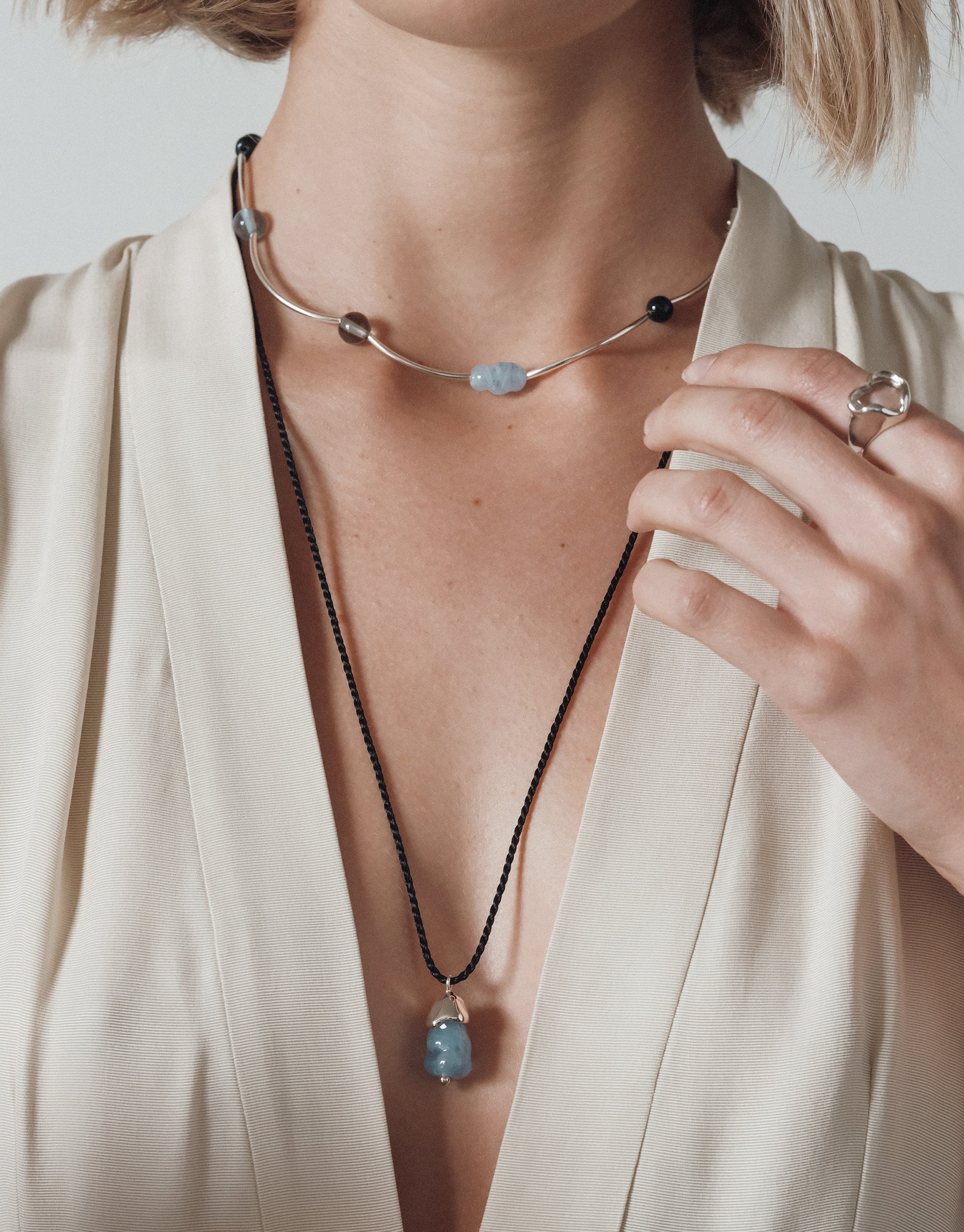 : A woman wears two layered necklaces—a silver choker with curved segments and small recycled glass beads in blue and smoky hues, and a long black cord necklace with a blue upcycled glass pendant. She wears a cream v-neck top and a sculptural silver ring.