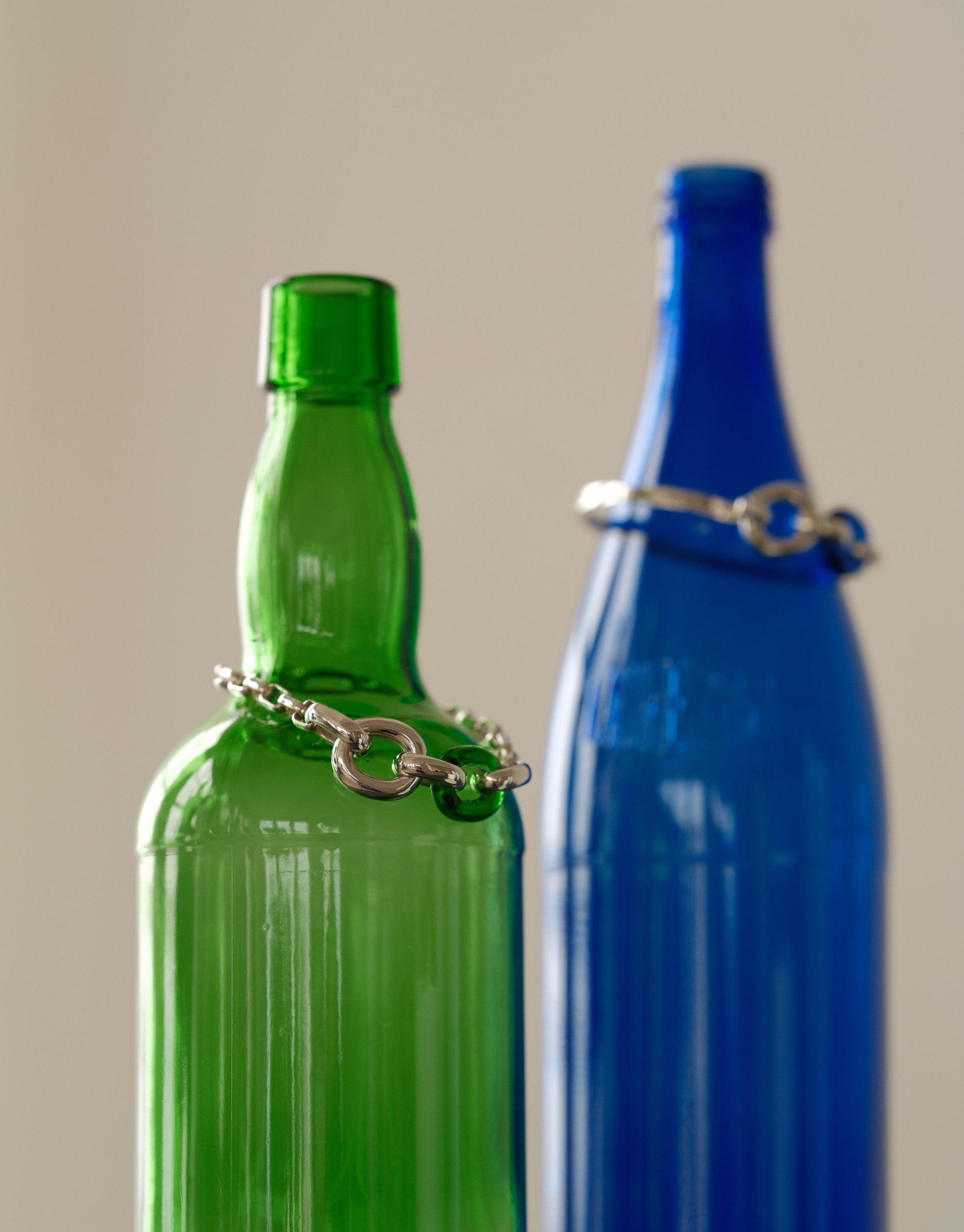 Loop Hook Bracelet displayed on upcycled green and blue glass bottles, showcasing sustainable jewelry design. The silver bracelets wrap elegantly around the bottle necks, emphasizing the unity and continuity of the In The Loop collection. Eco-friendly fashion statement by CLED.
