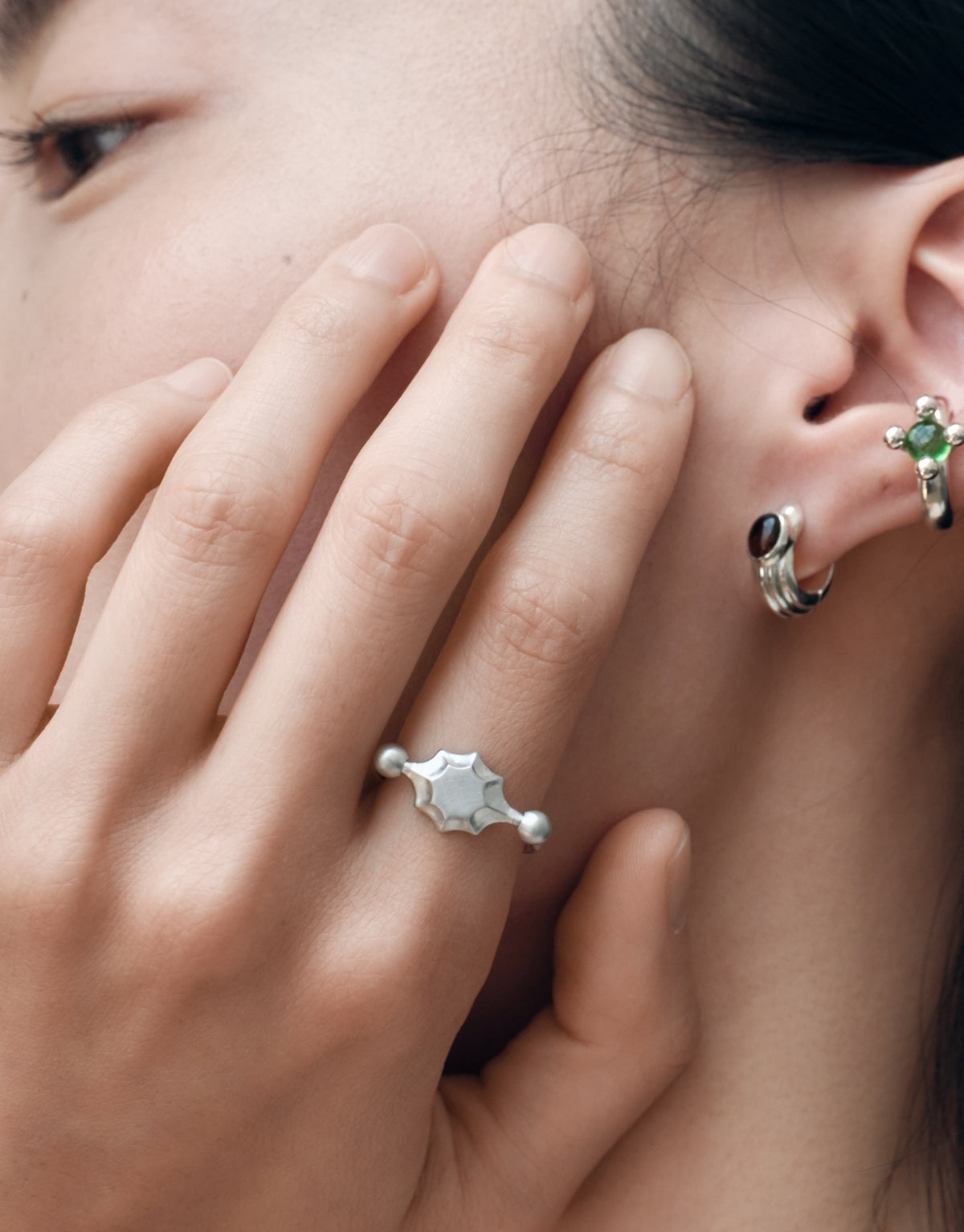 A close-up lifestyle shot of a model's hand and ear, showcasing a silver ring with a starburst design on the ring finger and a silver ear cuff with a green gemstone on the ear lobe.
