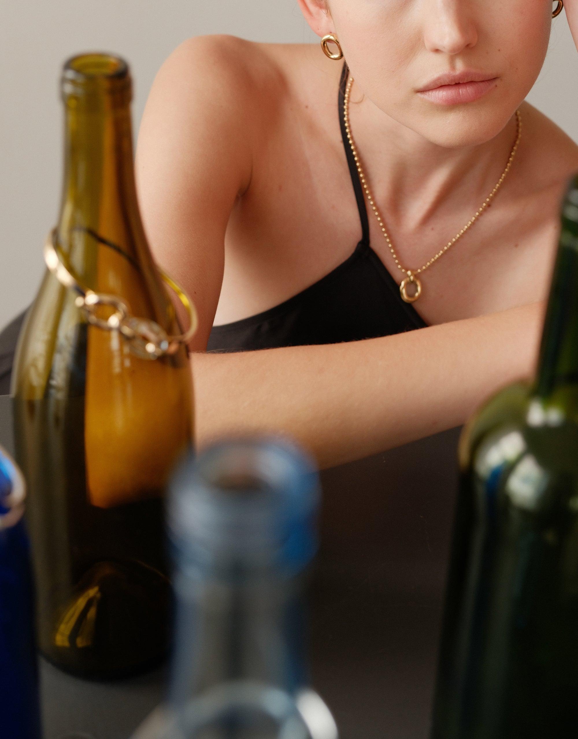 Elegant Core Loop Necklace on model, surrounded by upcycled glass bottles, showcasing sustainable jewelry design. Ball chain adds delicate touch to neckline. Represents unity and connection in CLED's In The Loop collection. Core Loop Necklace | Ball Chain by CLED.