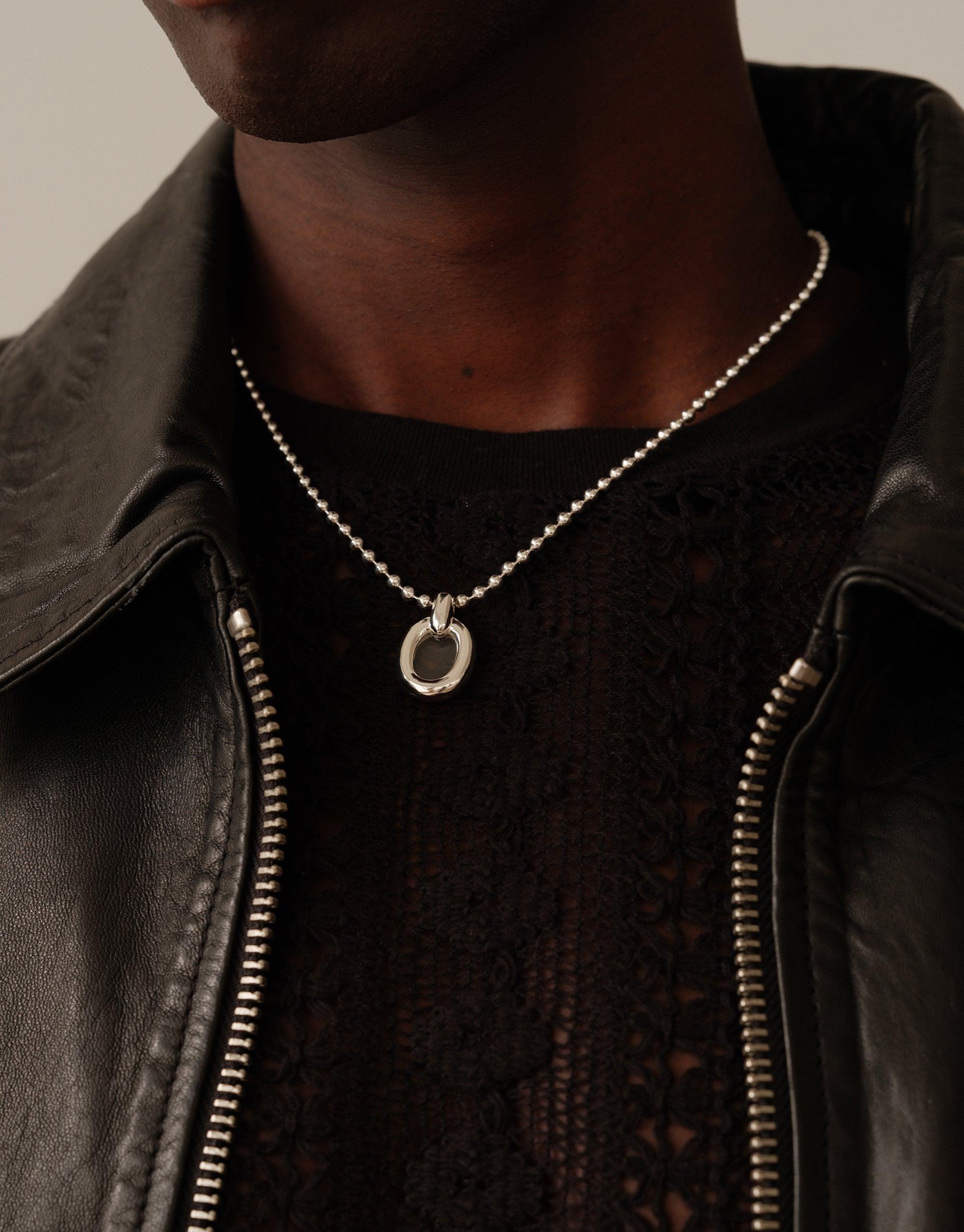 Close-up of a person wearing a Core Loop Necklace with ball chain. The silver pendant hangs elegantly against a dark top, complementing a leather jacket. The necklace symbolizes unity and upcycling, embodying CLED's sustainable ethos. Core Loop Necklace | Ball Chain by CLED.