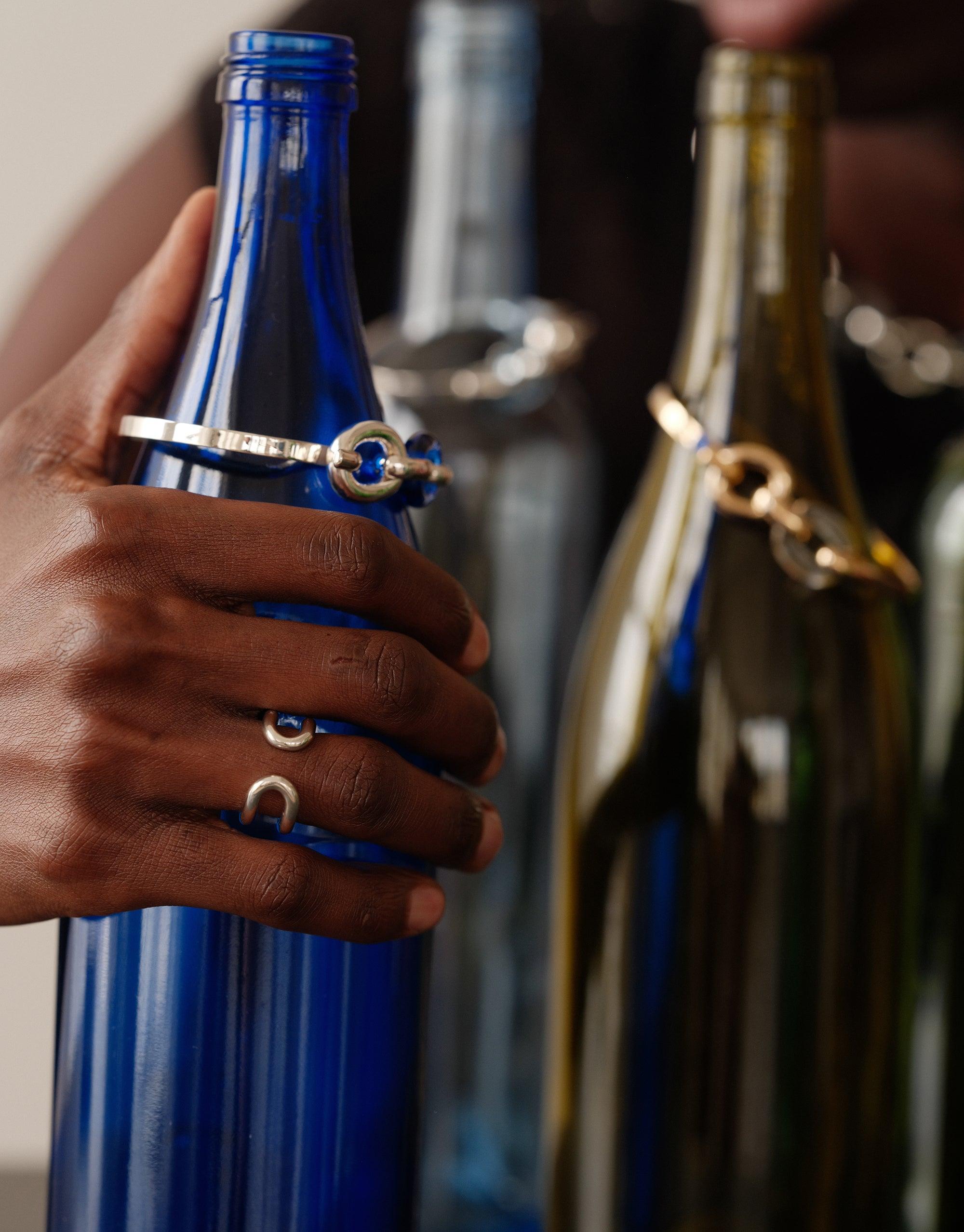 Hand holding blue glass bottle, showcasing Infinite Loop Ring and other upcycled jewelry pieces from the Loop Collection. Sustainable fashion accessories displayed on colorful repurposed bottles, representing unity and environmental consciousness. Infinite Loop Ring by CLED.
