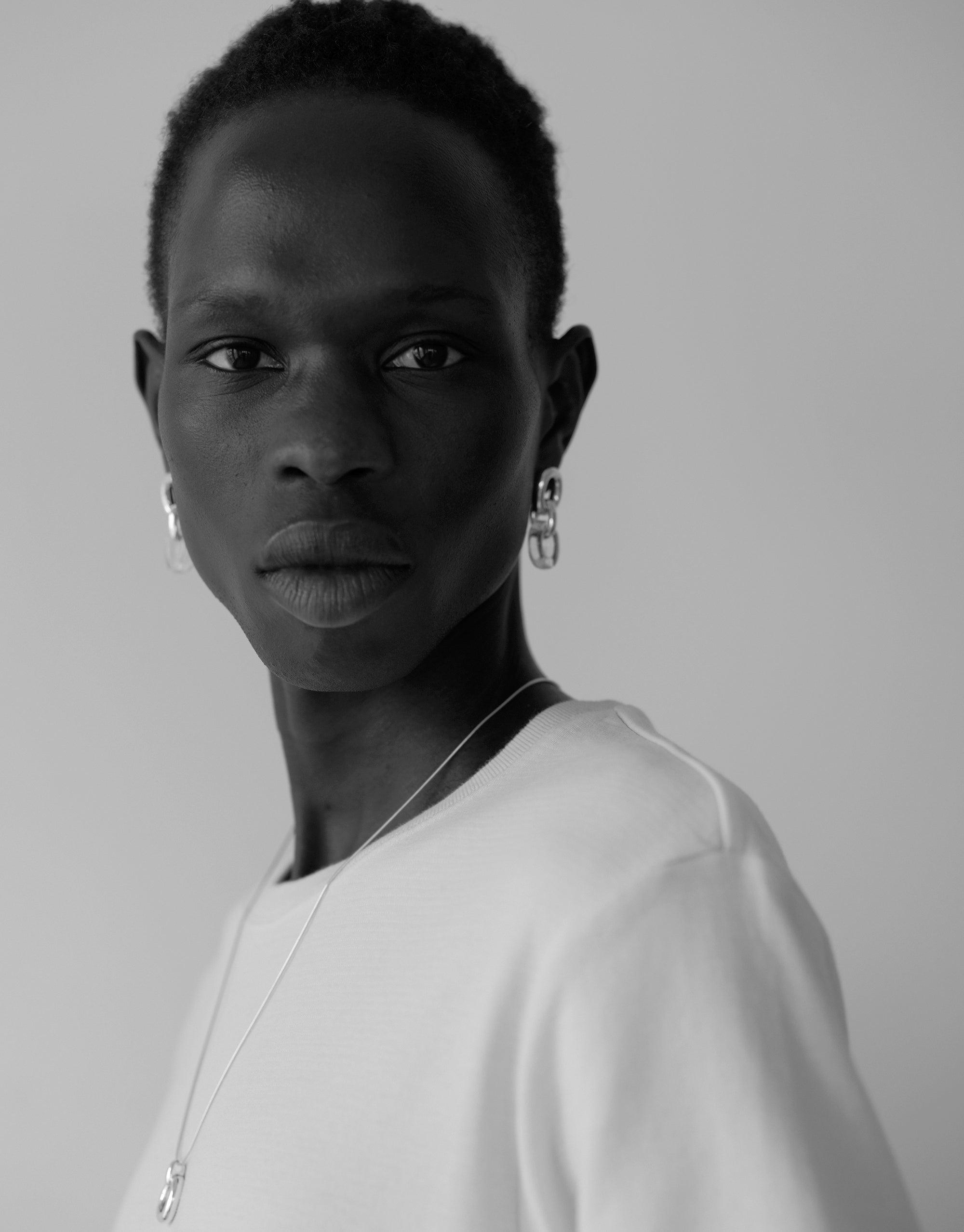 Black and white portrait showcasing Connected Loop Earrings, emphasizing simplicity and elegance. Model wearing white top and delicate necklace, highlighting the earrings' unique design. Represents unity and upcycling ethos of the Loop Collection. CLED Connected Loop Earrings.