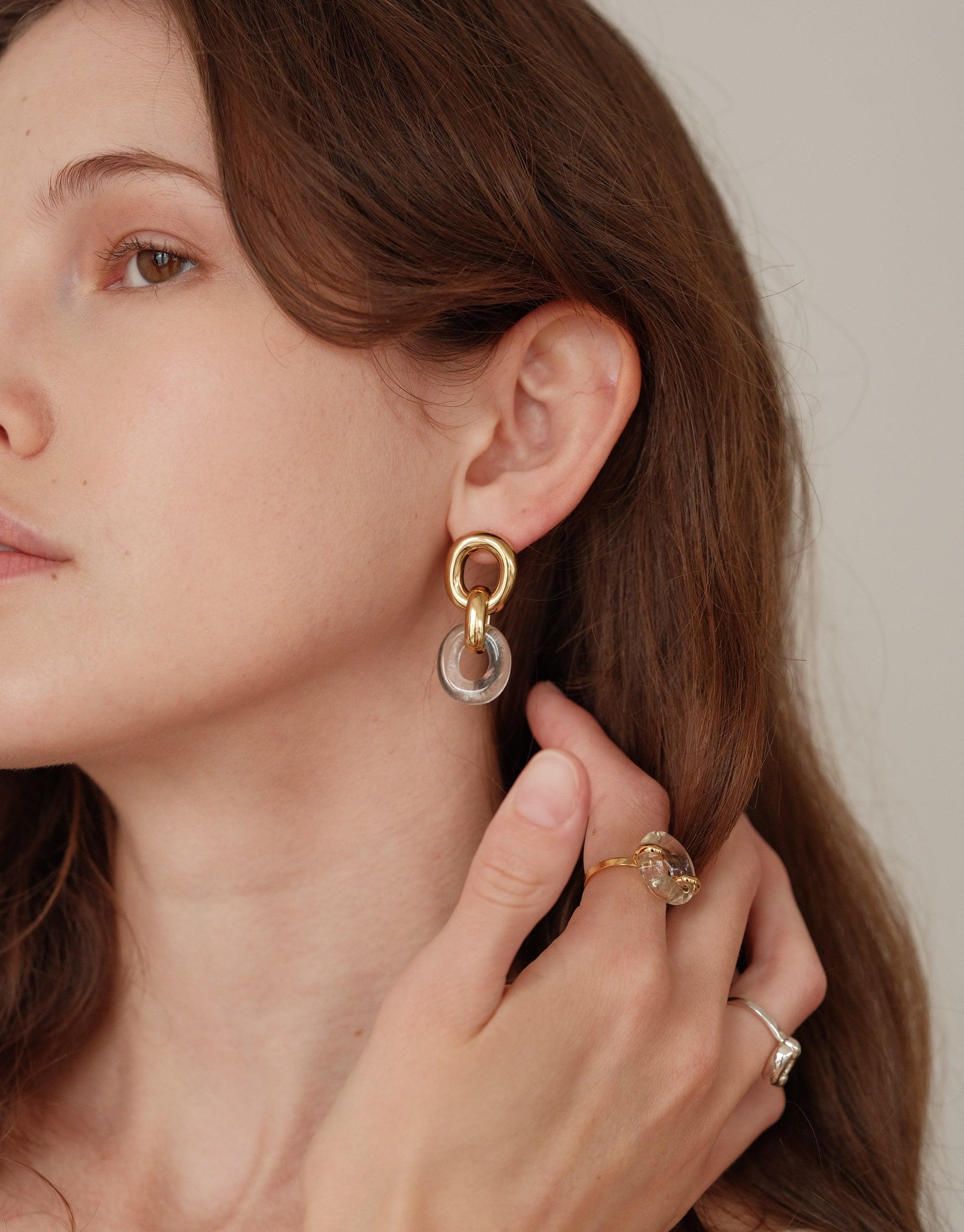 Elegant Connected Loop Earrings showcased on model, featuring interlocking gold and clear circular elements. Symbolizes unity and upcycling, part of the In The Loop collection. Paired with complementary rings for a complete look. Sustainable jewelry by CLED. Connected Loop Earrings.