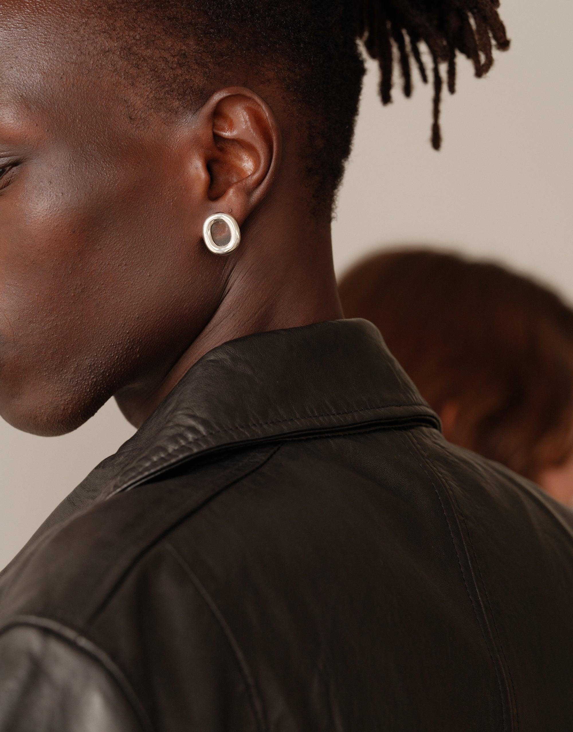 Close-up profile view of a person wearing large circular white earrings, showcasing the Core Loop Earrings | Large. The earrings contrast beautifully against dark skin and a leather jacket, emphasizing their elegant, minimalist design. CLED