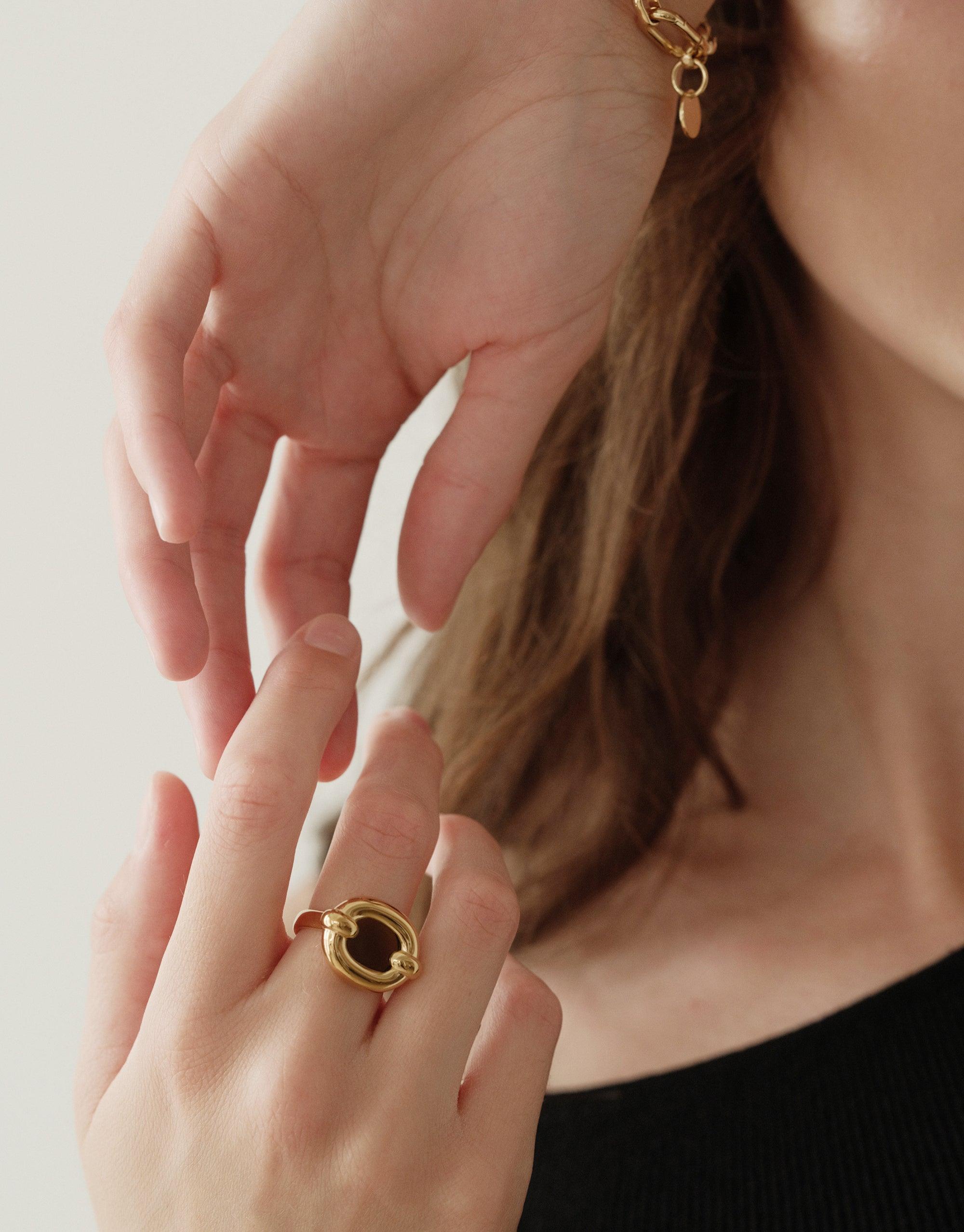 Close-up of a hand wearing the Core Loop Ring | Large, a gold-toned metal ring with a circular design featuring a mirror-like center. The ring is displayed on a person's finger against a light background, showcasing its unique reflective properties. CLED